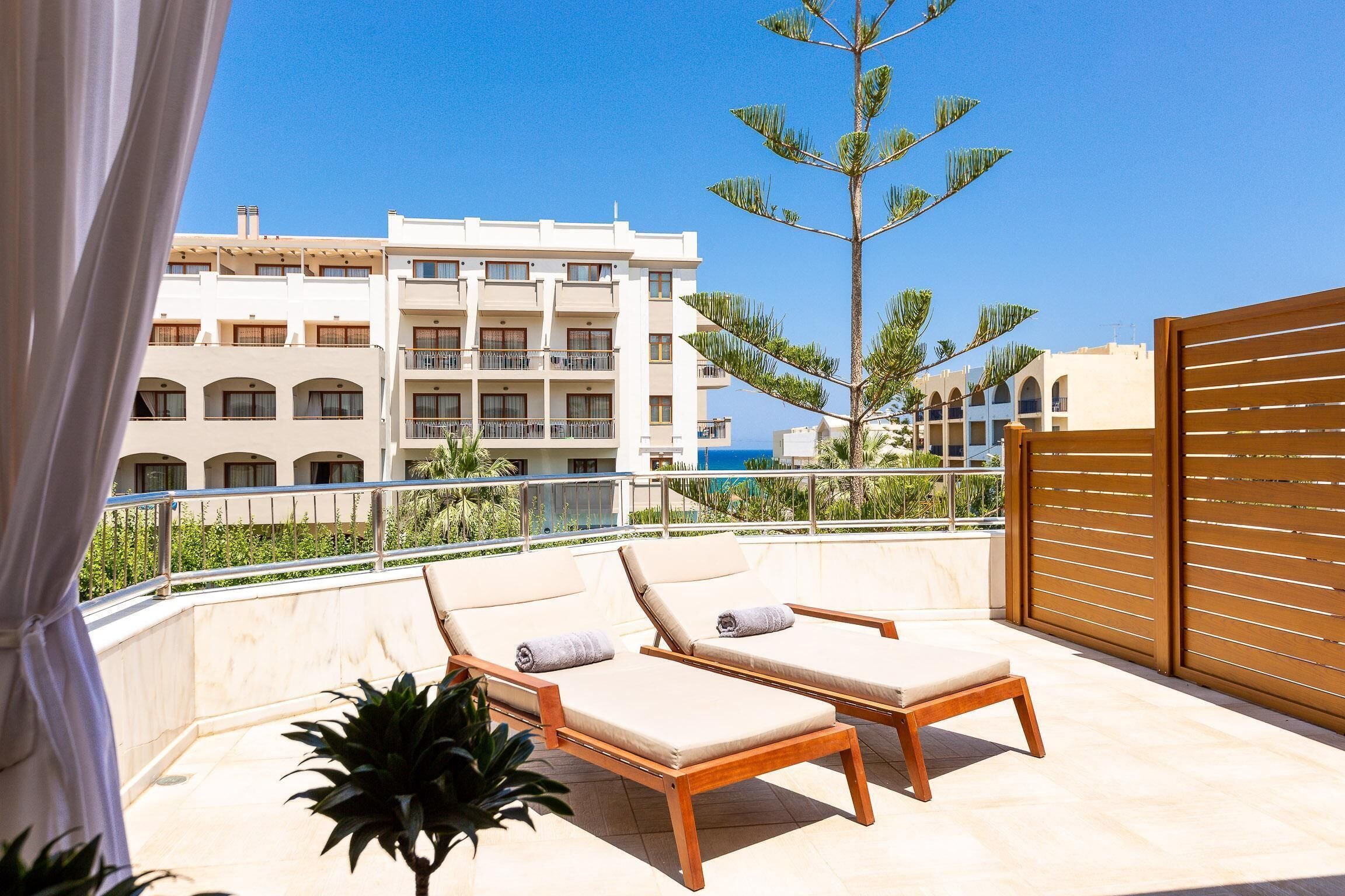 Un balcón con dos sillones de madera, una planta en maceta y vistas de un edificio con varios balcones y una palmera.