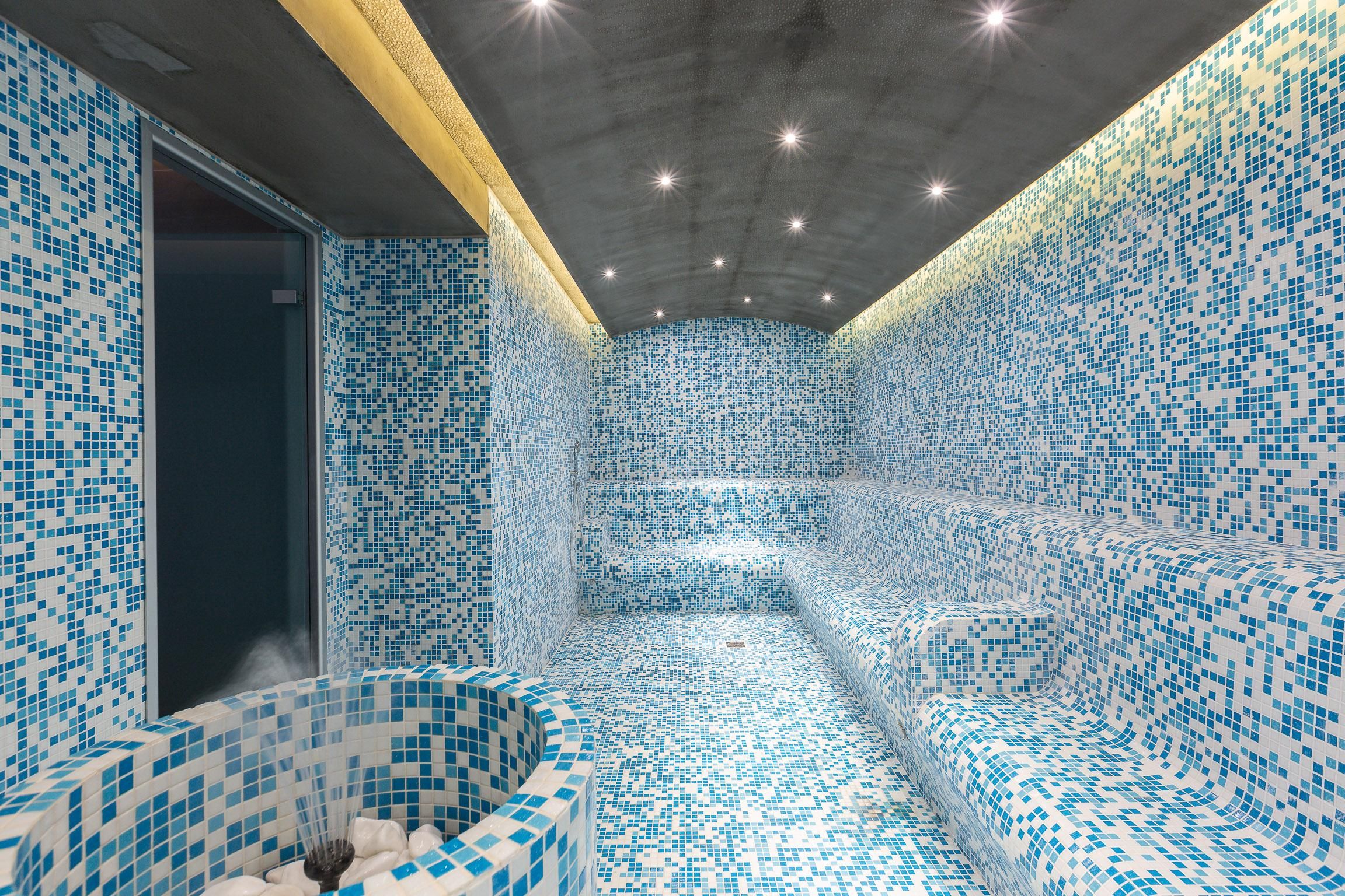 Un cuarto de spa con piso y paredes de azulejos, un área de ducha y una zona de estar con un banco.