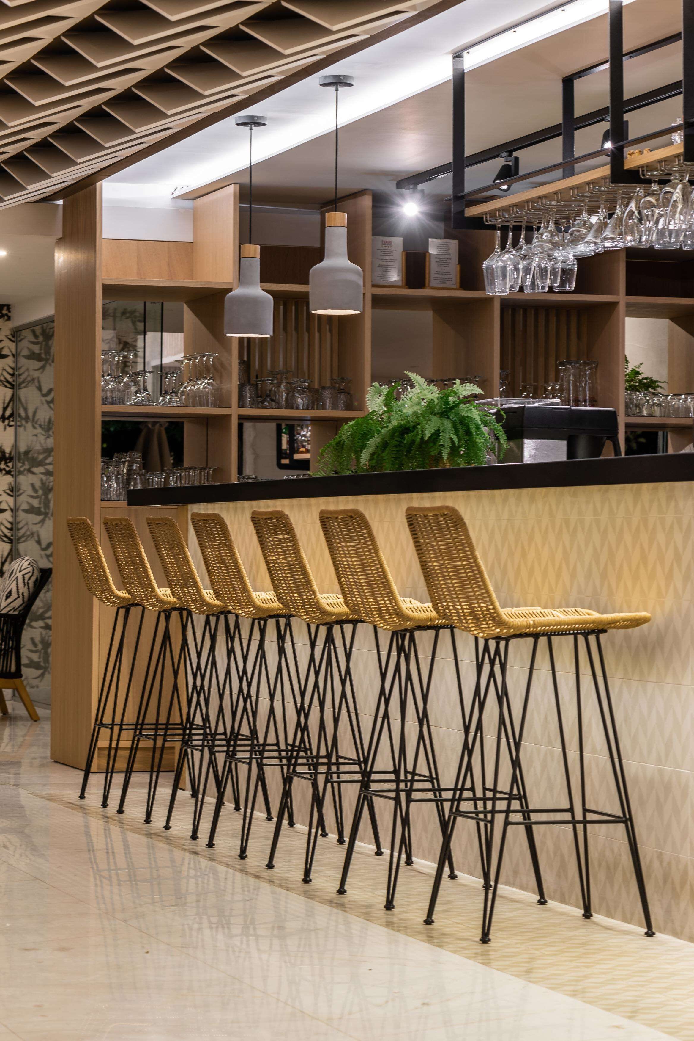 A bar area with wicker bar stools, hanging light fixtures, and shelves with glasses and bottles.