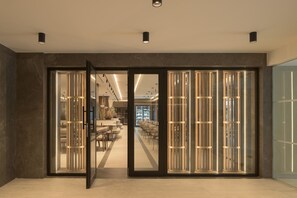 A modern interior with a glass door entrance, a seating area with tables and chairs, and a visible bar area in the background. - Theartemis Palace (Rethymno)