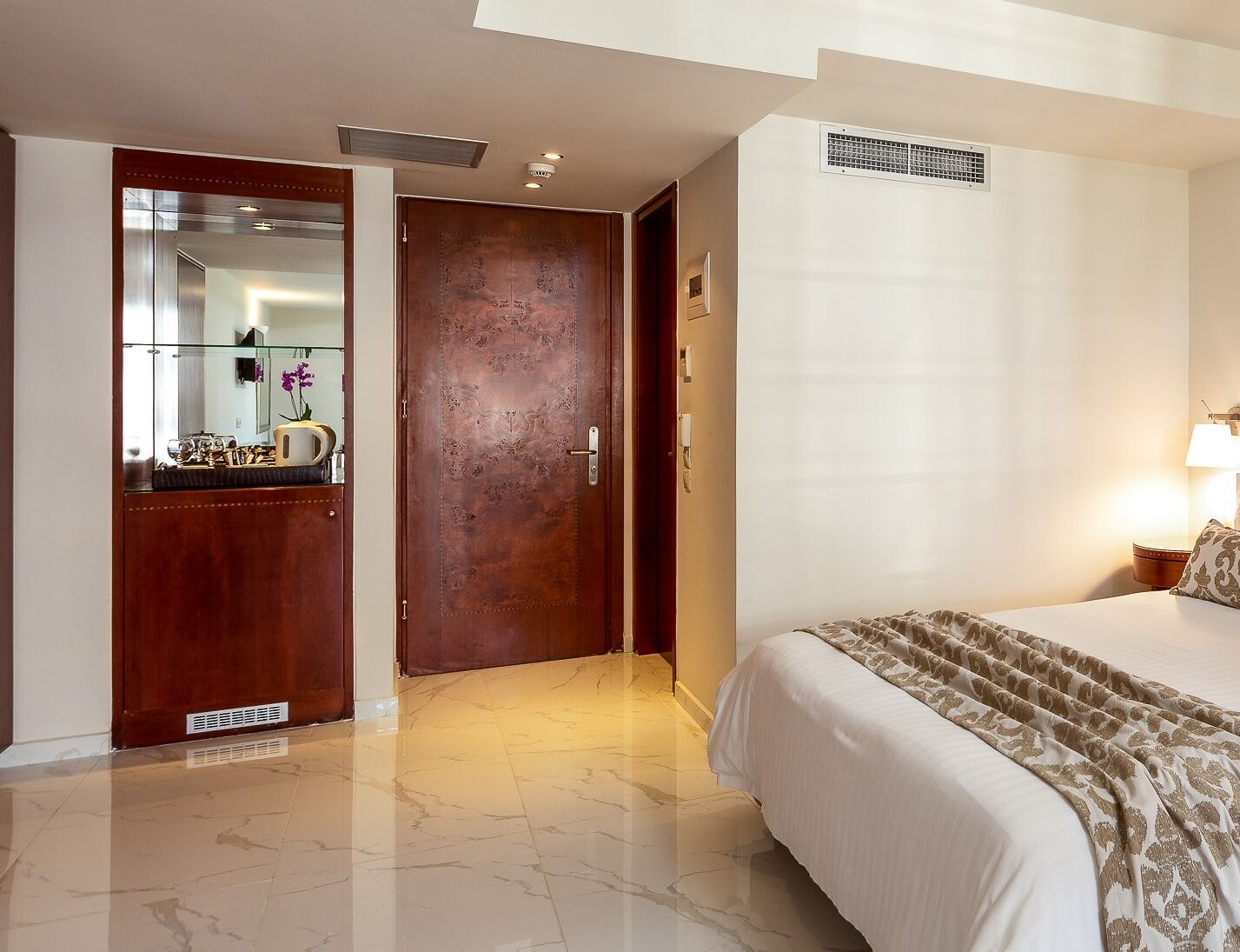 A hotel room with a bed, a wooden wardrobe, a mirror, and a bedside table with a lamp.