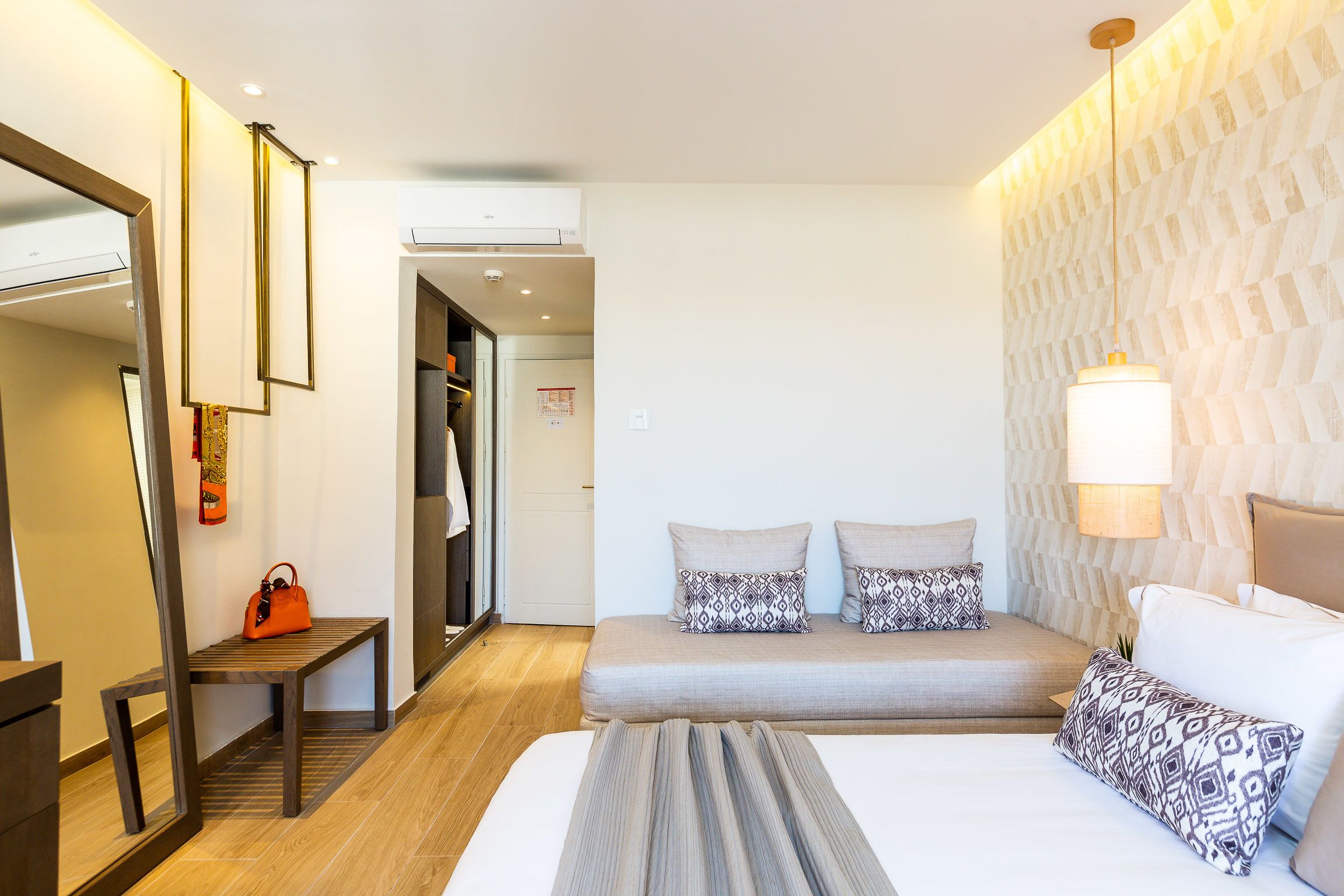 Una habitación de hotel con cama, mesita de noche y un banco con un bolso de mano.