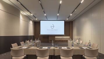 Un salón de conferencias con una larga mesa, sillas y una pantalla de proyector mostrando 'Theartemis Palace RETHYMO'.