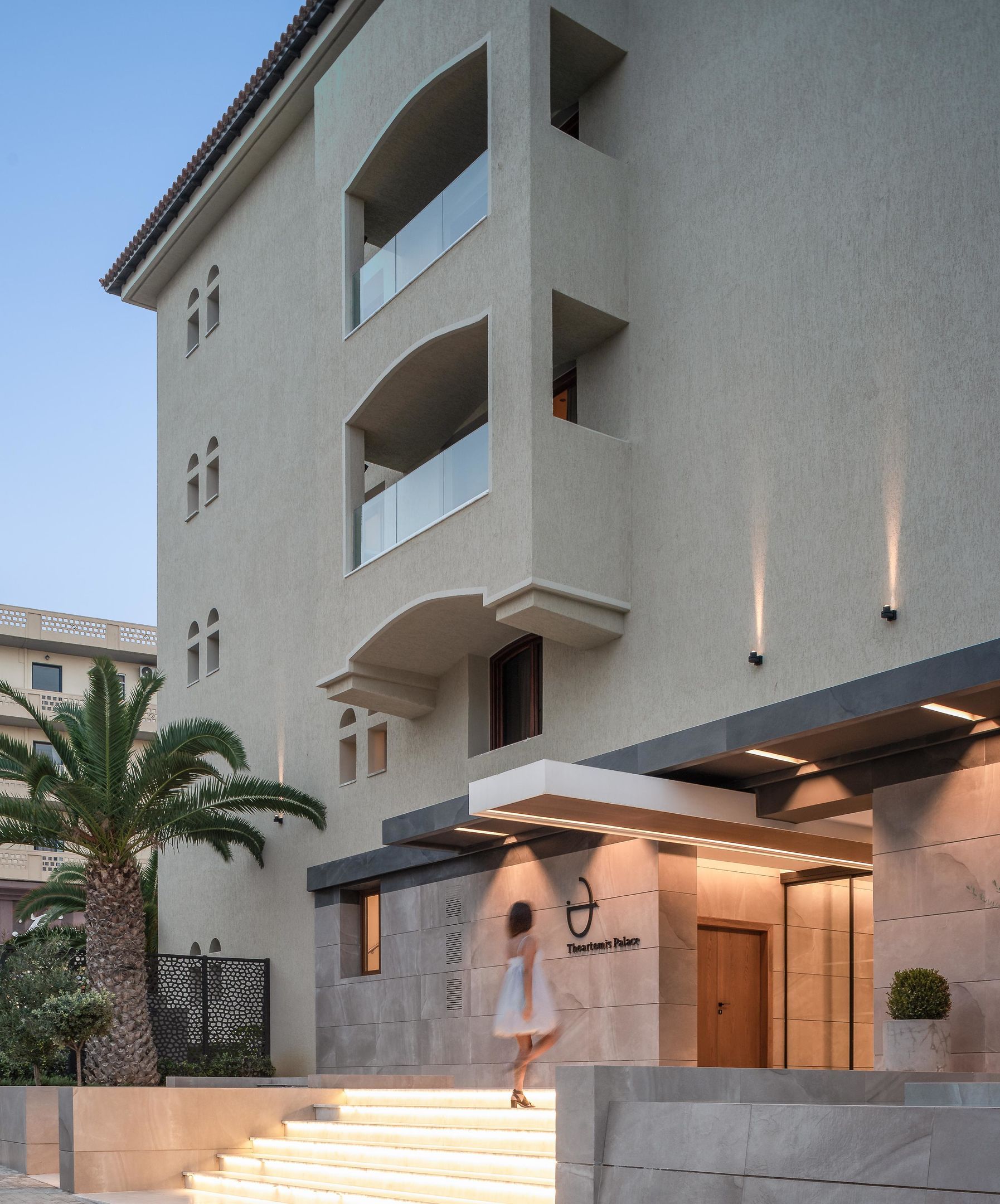 A multi-story building with a modern architectural design, featuring a prominent entrance with steps leading up to a door, and a sign indicating the name of the property.