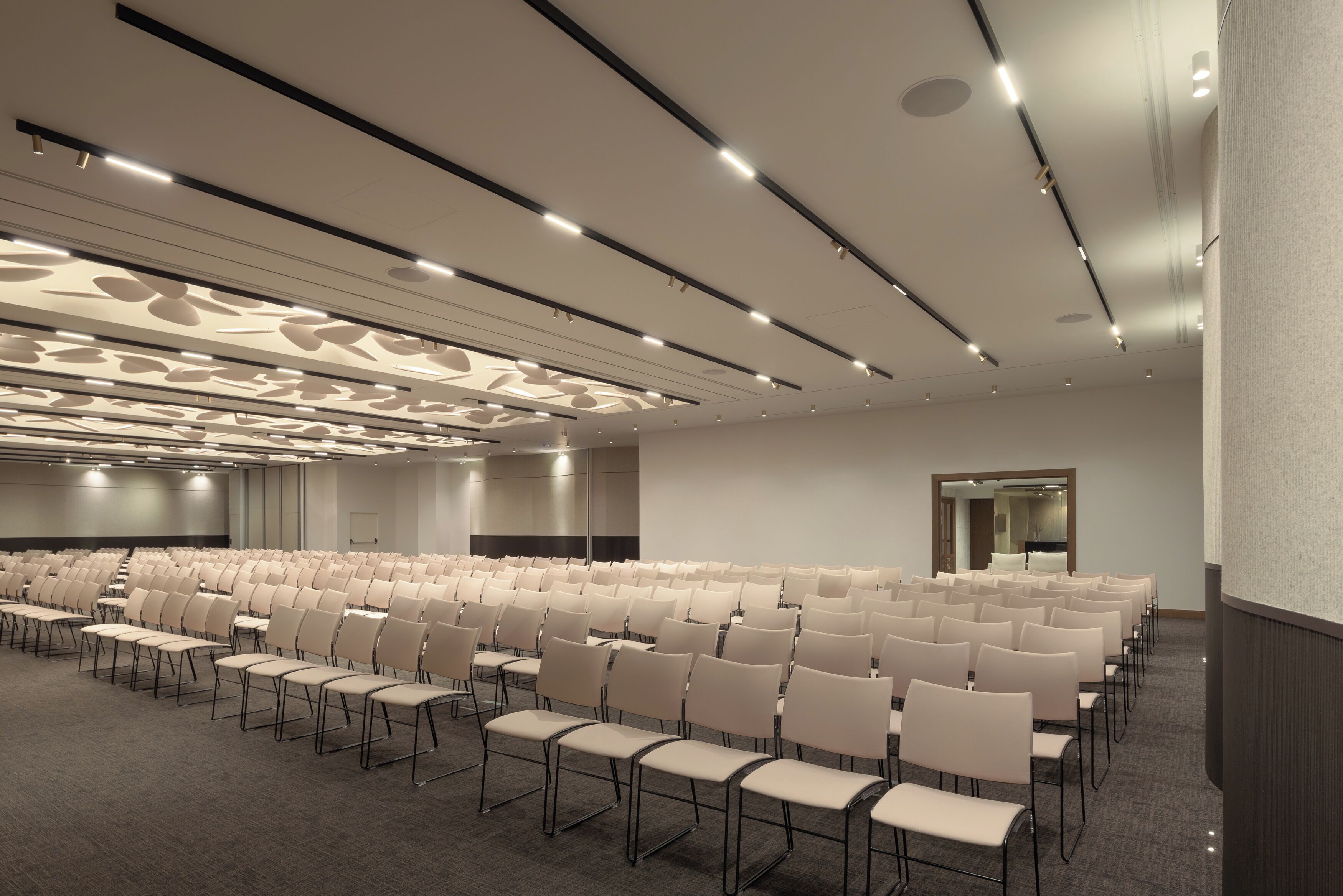 Une salle de conférence avec des rangées de chaises, un sol recouvert de moquette et un plafond au design moderne doté d’un éclairage encastré.