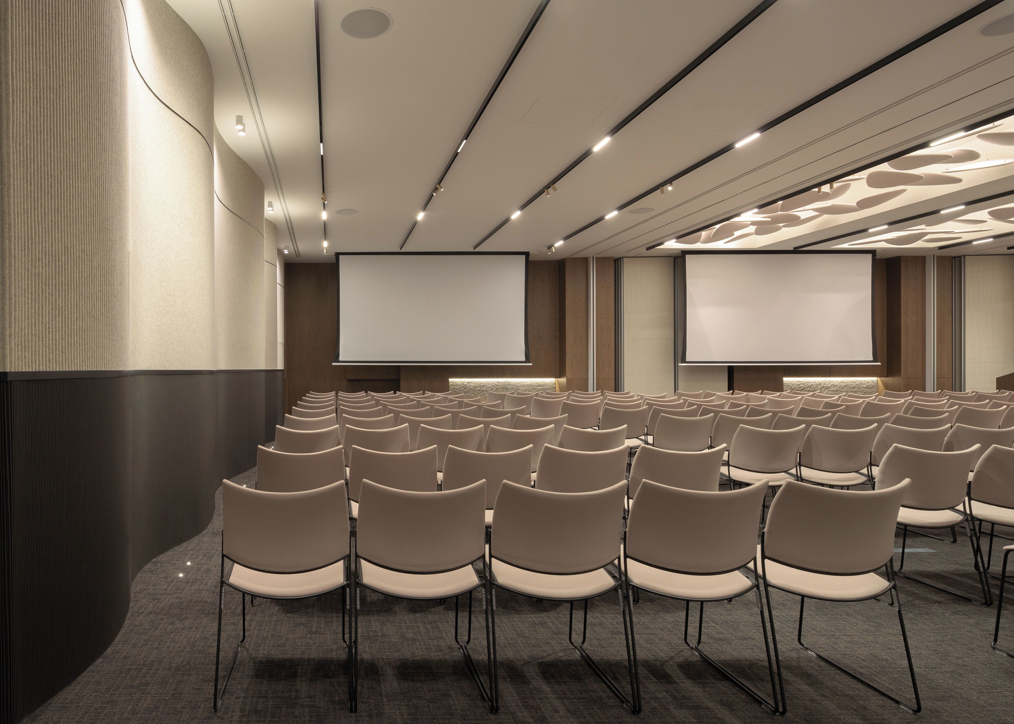 Un salón de conferencias con filas de sillas, una pantalla grande y piso alfombrado.