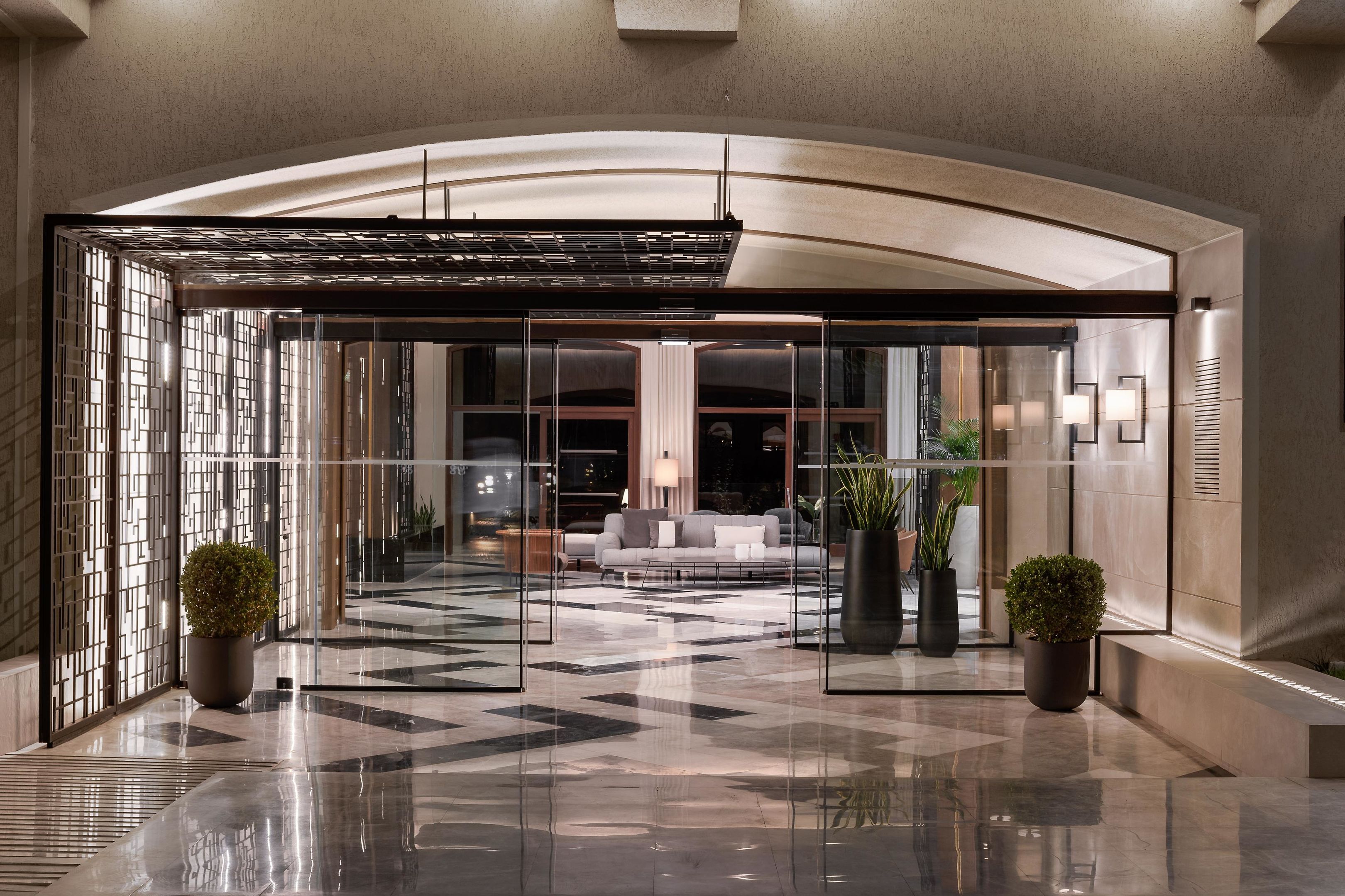 A modern lobby with a glass entrance, a seating area with a sofa, and potted plants.