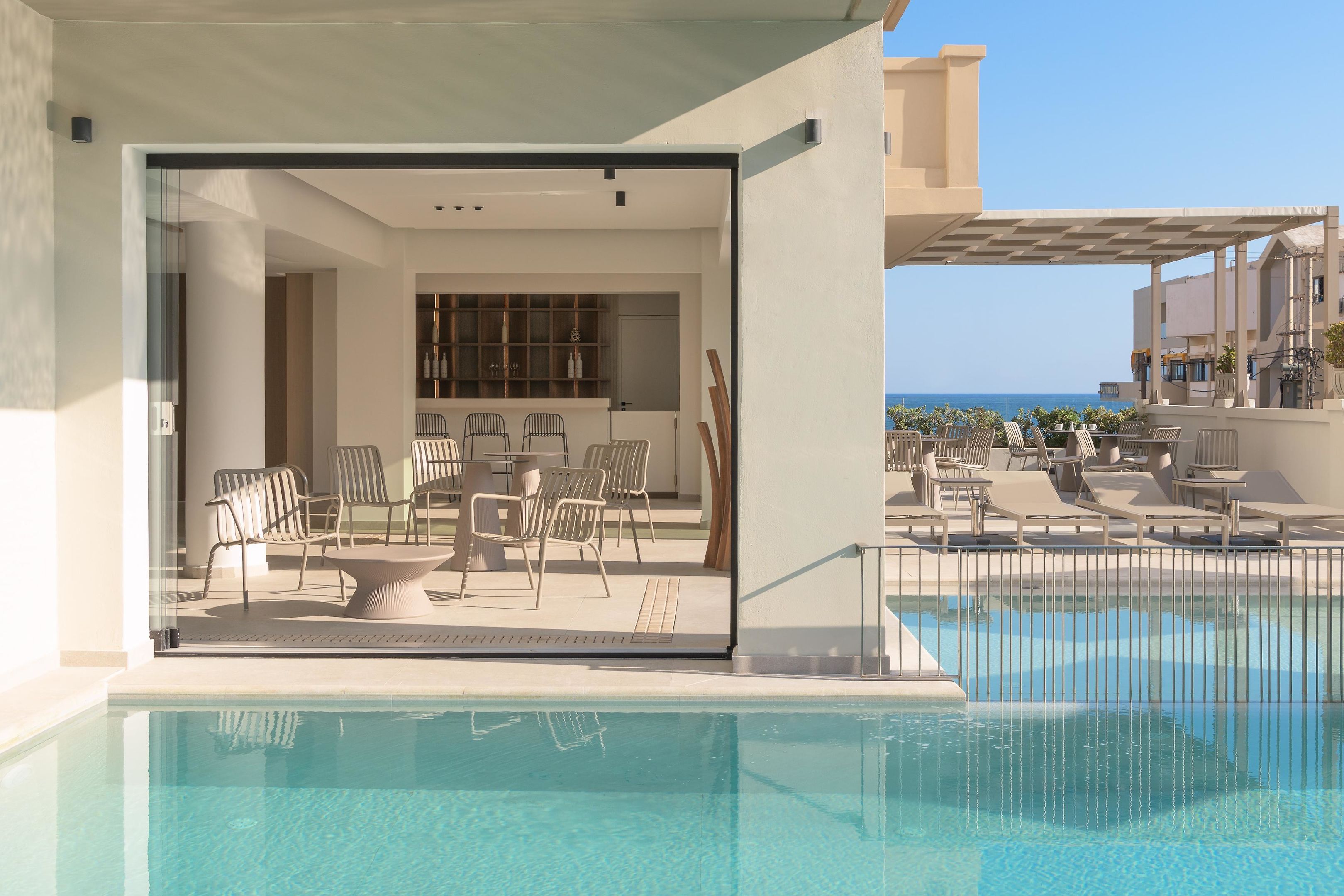 A modern poolside area with a clear view of the ocean, a dining area with chairs and a table, and a bar area with shelves and bottles.