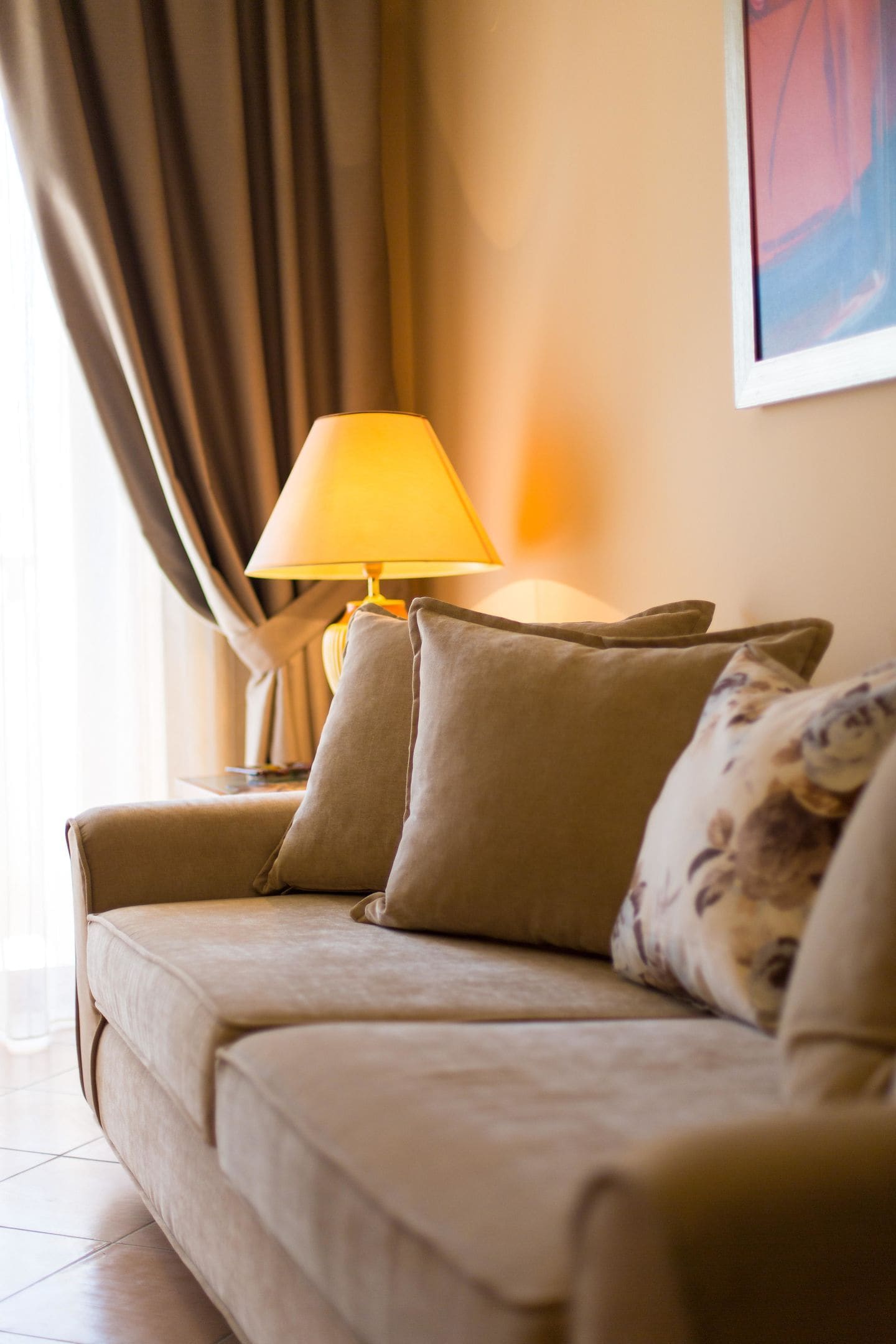 A beige sofa with three cushions in a room with a lamp and curtains.