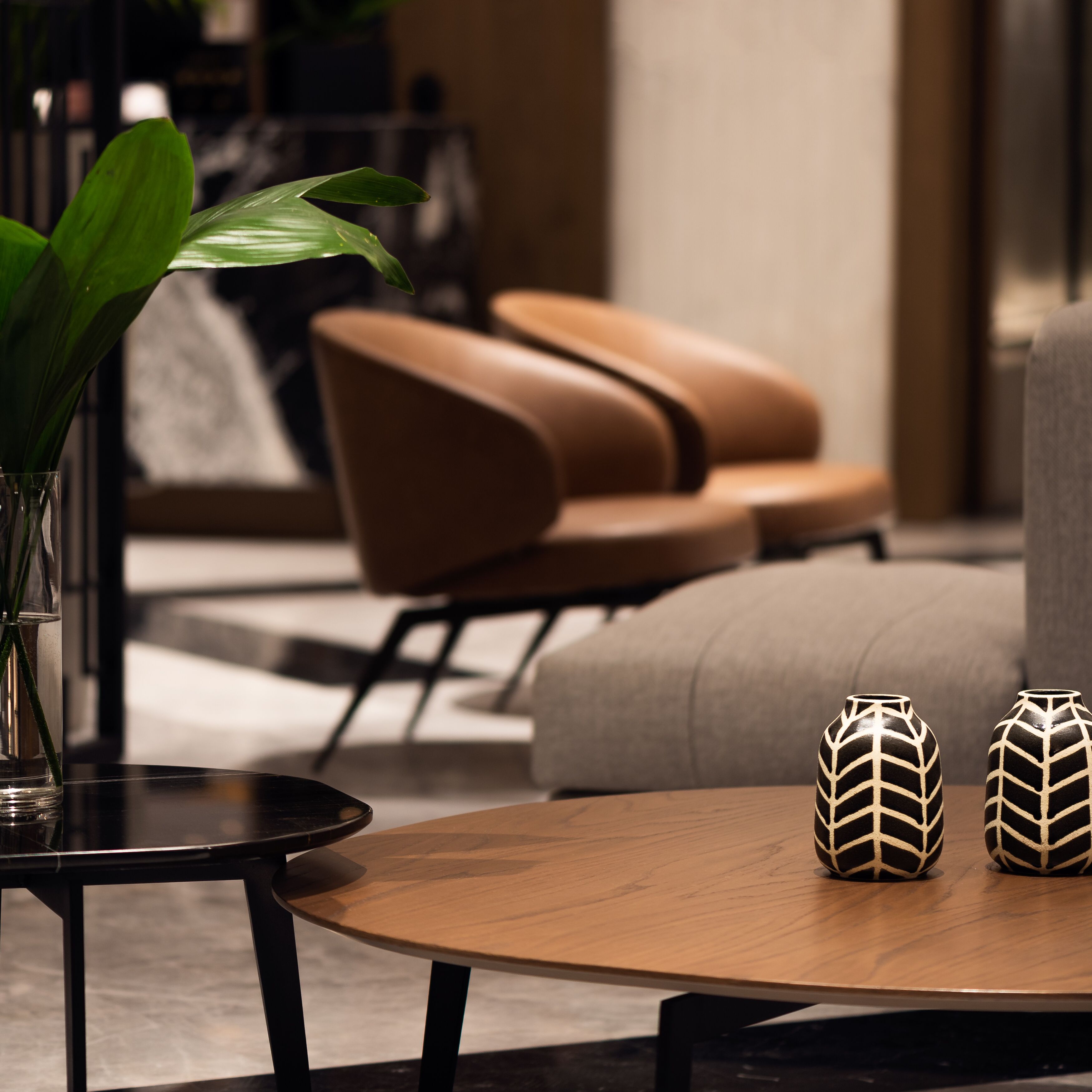 Een moderne woonkamer met een ronde houten salontafel, twee decoratieve vazen, een potplant en een bruine leren bank.