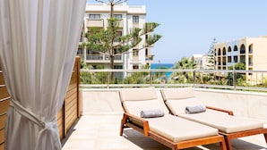 A balcony with two sun loungers, a white curtain, and a view of a building and the sea.