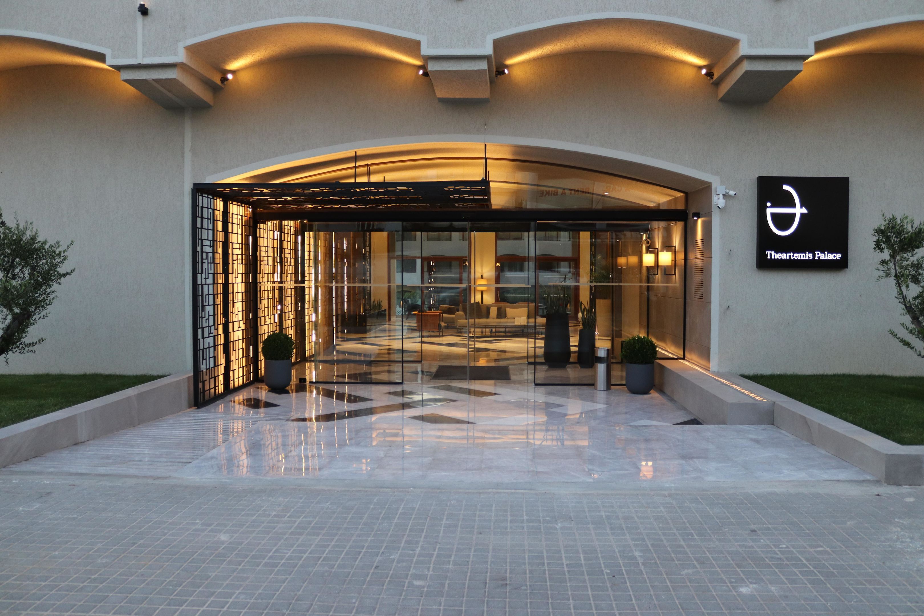 Entrada de hotel con fachada de vidrio, un letrero que indica Theartemis Palace y piso de baldosas.