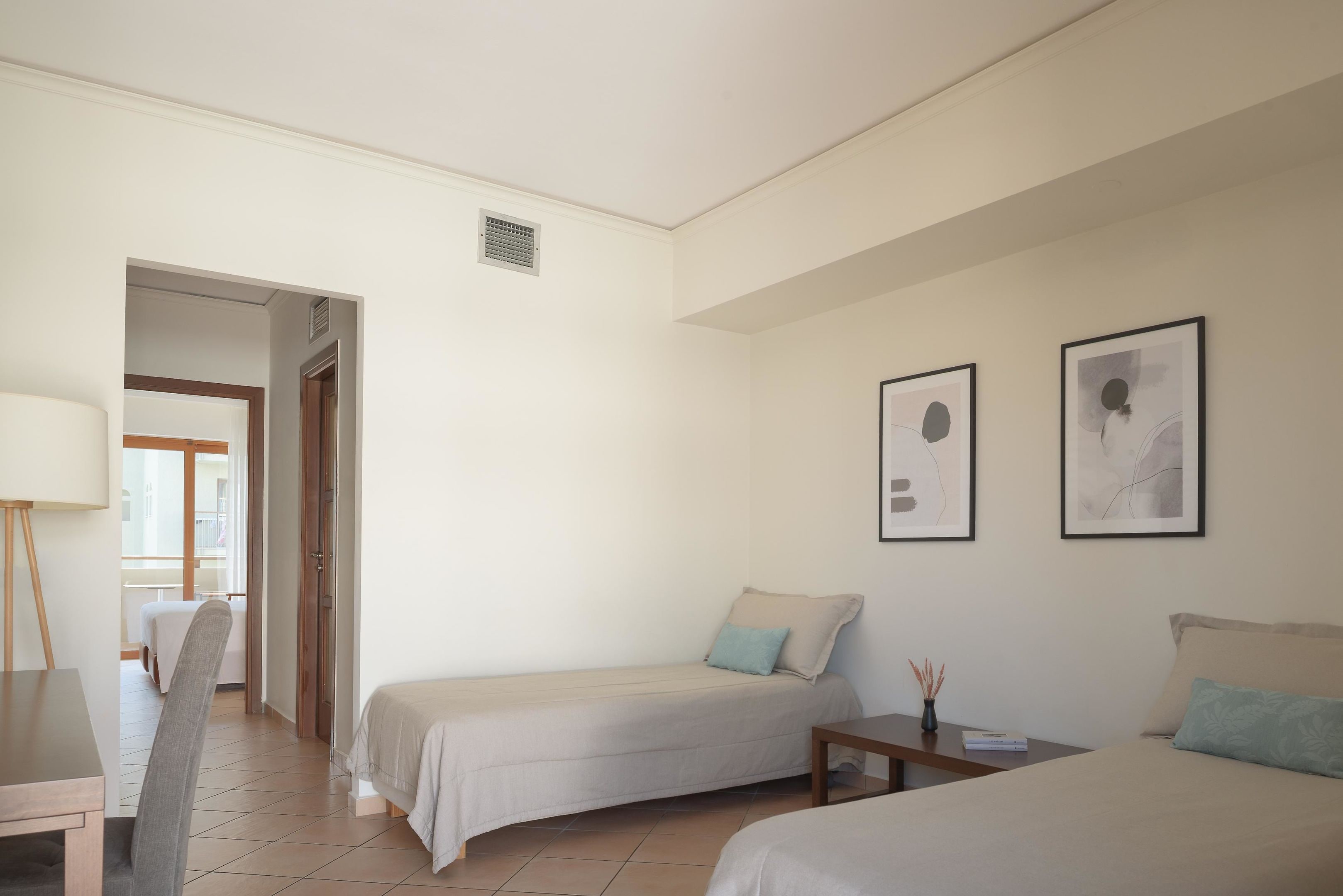 A hotel room with two beds, a desk, a chair, and a door leading to a balcony.