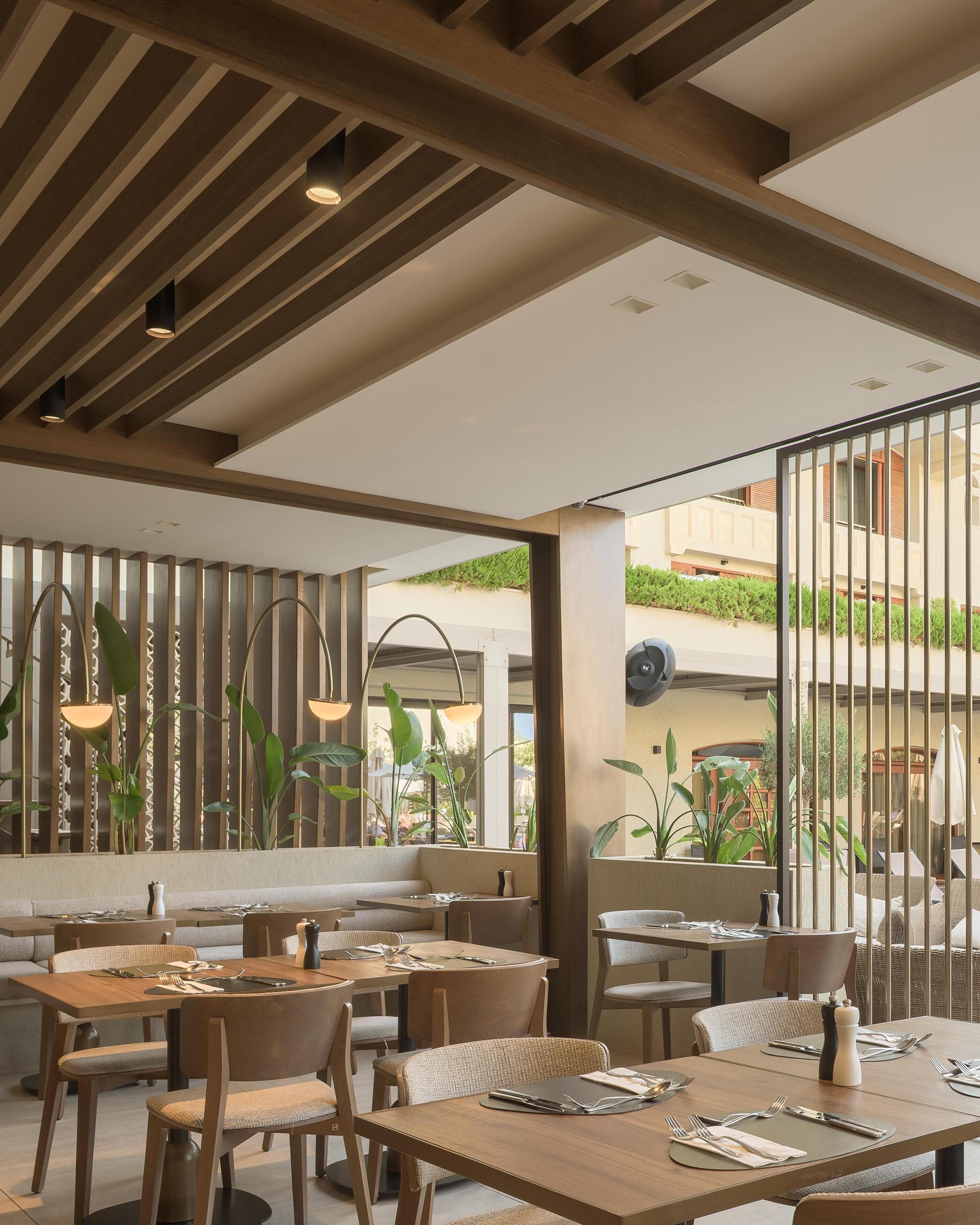 Un restaurant avec des tables et des chaises en bois, de grandes fenêtres et des plantes d’intérieur.