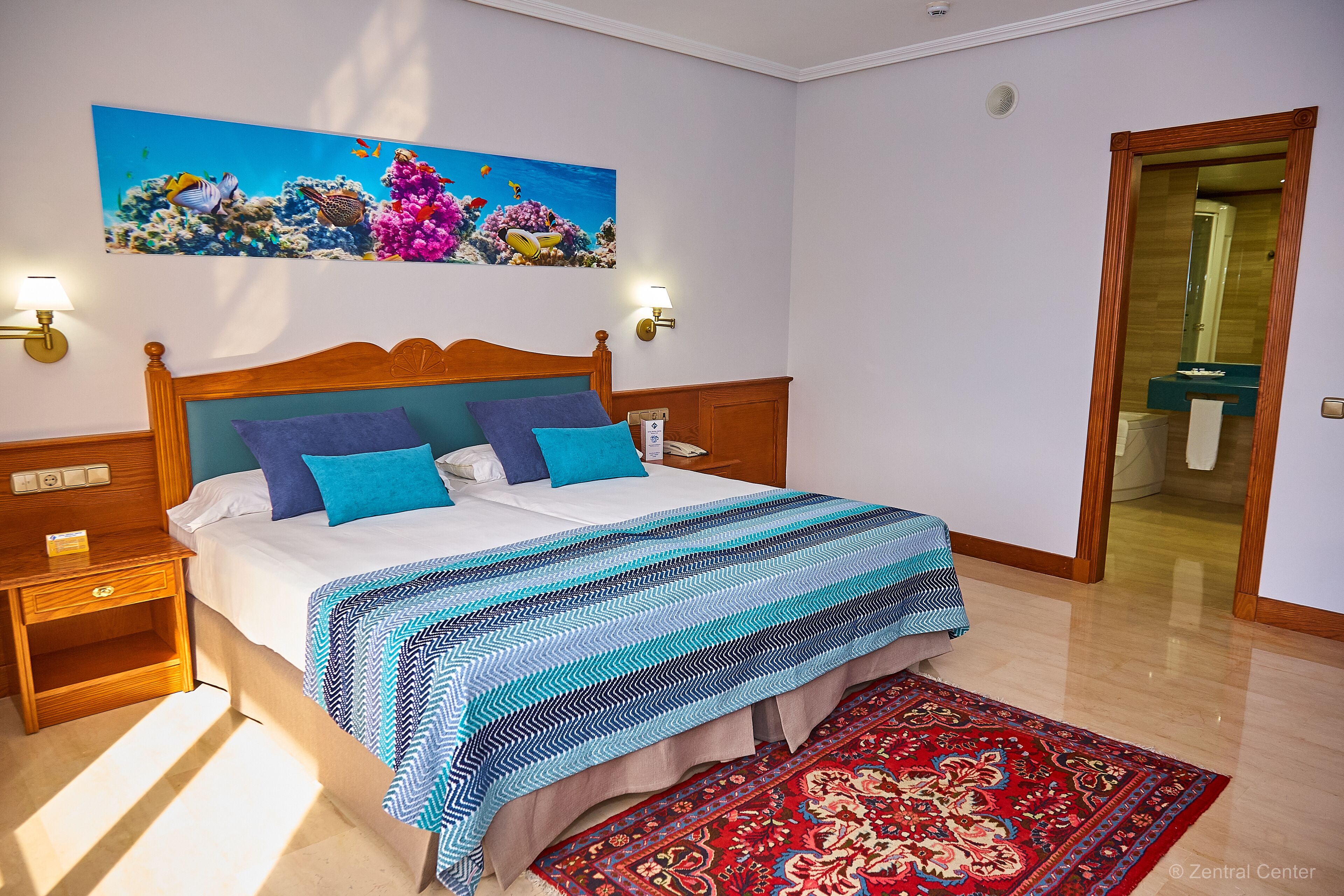 Une chambre d’hôtel avec un lit, une table de chevet, une lampe fixée au mur, un tableau représentant un paysage sous-marin et un tapis rouge à motifs.