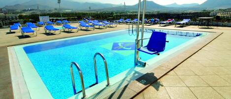 A rooftop swimming pool with blue lounge chairs and a view of mountains.