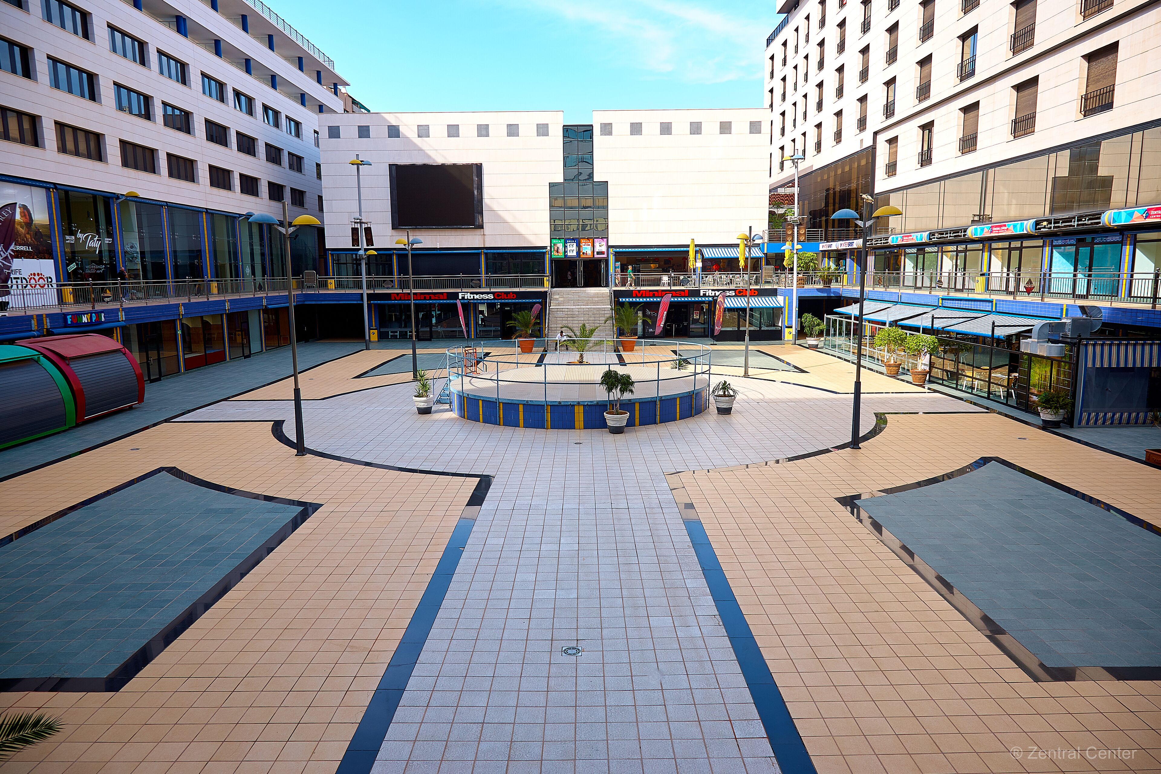 A modern urban plaza with a central fountain, surrounded by commercial buildings with large windows and storefronts.