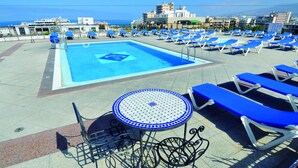 A rooftop pool area with blue lounge chairs and a small table, overlooking a cityscape and the sea. - Hotel Zentral Center (Arona)