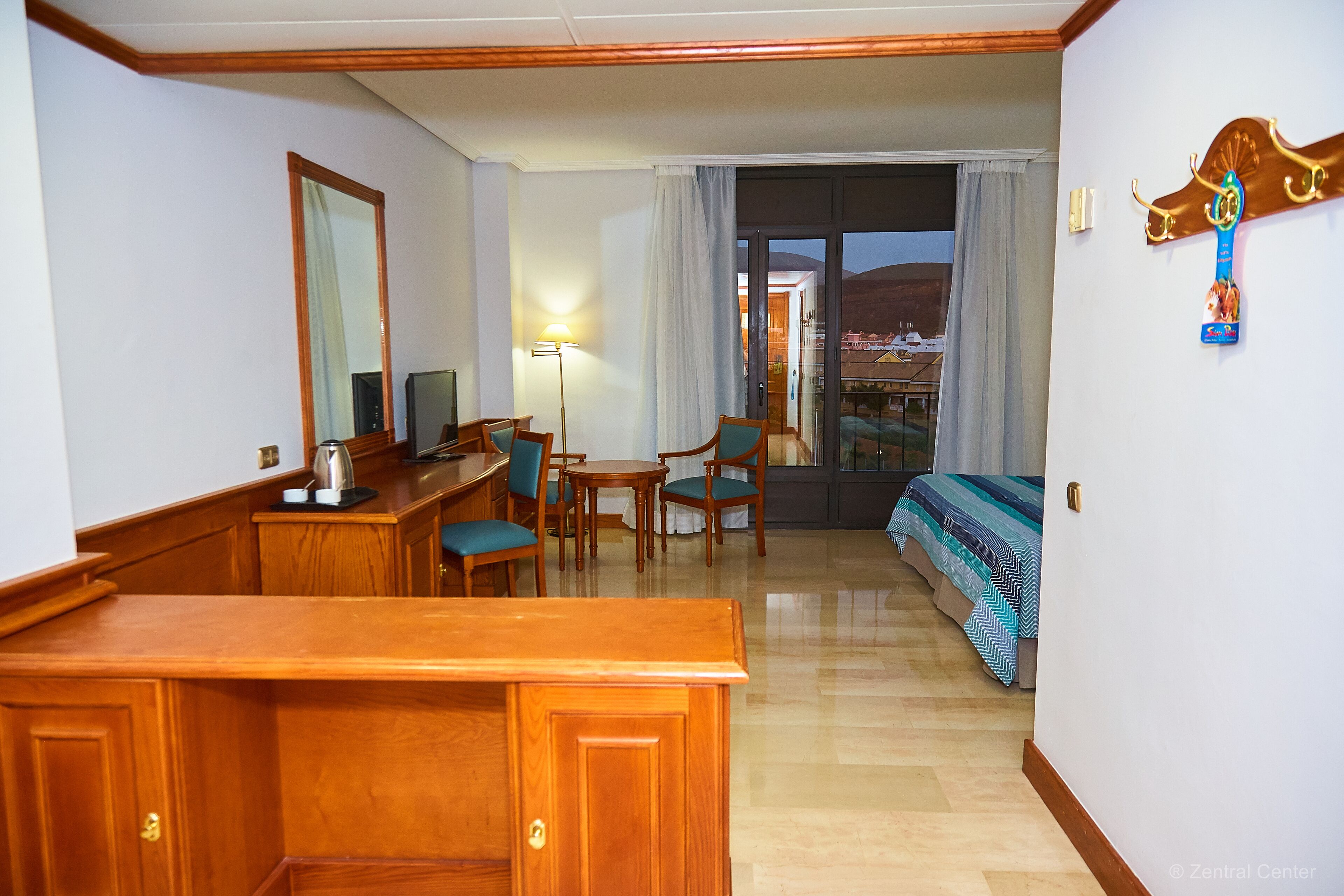 A hotel room with a wooden desk, two chairs, a bed with a striped bedspread, a large mirror, and a view of a mountainous landscape through the window.