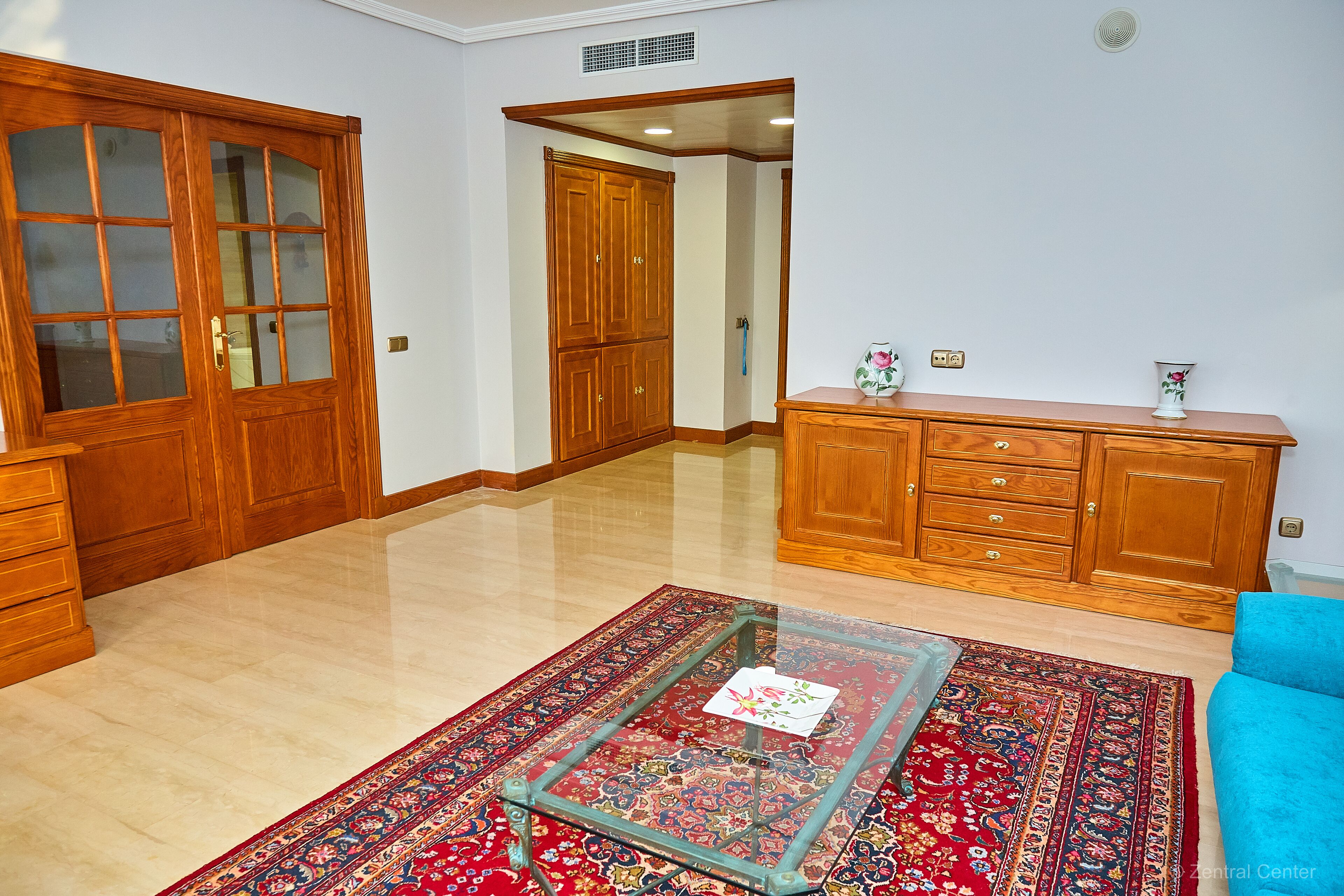 Un salón espacioso con un amplio armario de madera, una mesa de centro de cristal y un sofá azul.