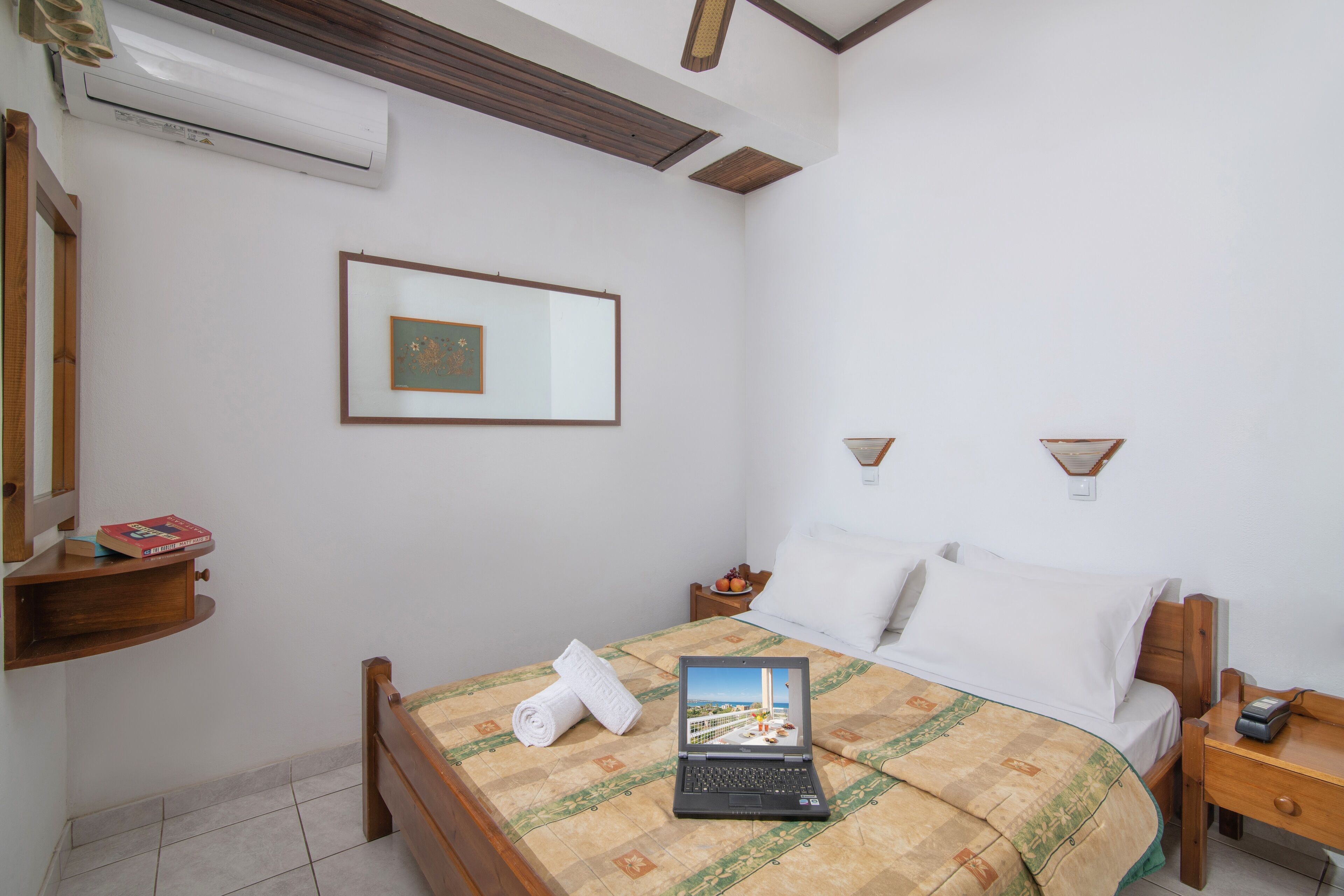 Een netjes ingerichte slaapkamer met een bed, een laptop, een nachtkastje, een wandairco, een lijst met een afbeelding en een plank met boeken.