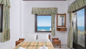 A hotel room with a bed, a window with a view of the sea, a wooden nightstand, and a small table with a chair.