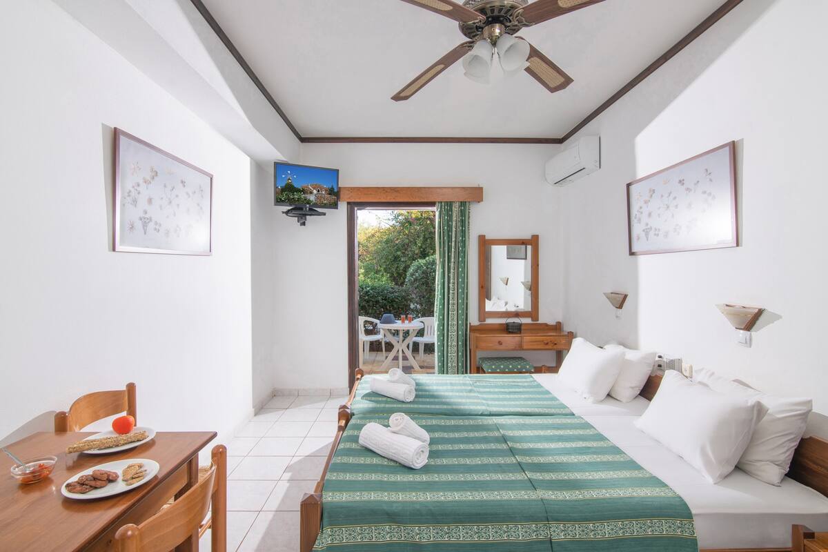 Habitación de hotel con cama, mesa de comedor, sillas, ventilador de techo y vistas al exterior.