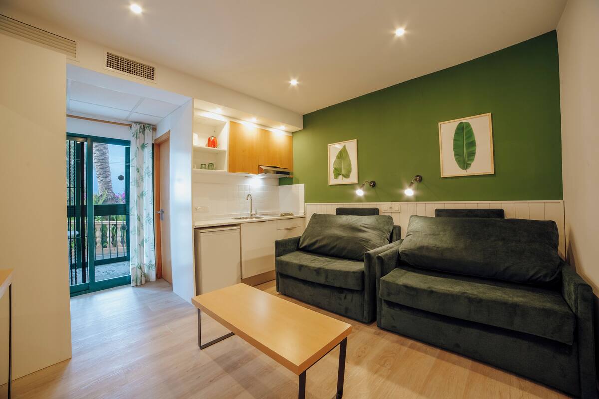 A modern living room with a wooden table, two green sofas, a kitchen area with white cabinets, and a green accent wall with leaf artwork.