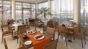 A dining area with multiple tables set for a meal, large windows with curtains, and a palm plant in the center. - Praia Sol (Vilamoura)