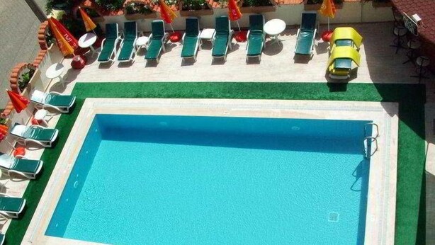 A rectangular swimming pool with a green border, surrounded by lounge chairs and umbrellas. - Melodi (Marmaris)