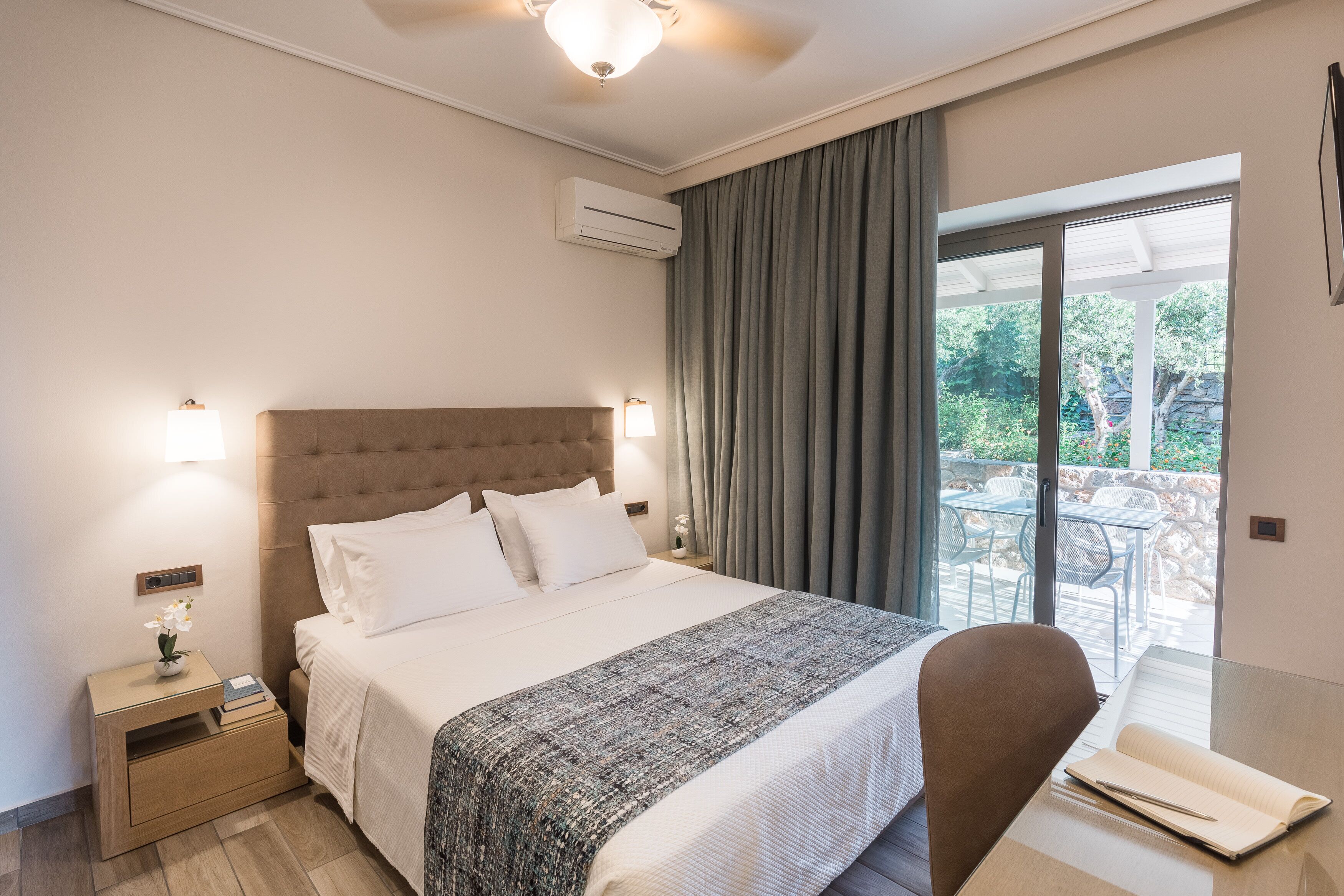A hotel room with a large bed, a nightstand with a lamp, a desk with a chair, and a view of the outdoors through a sliding glass door.