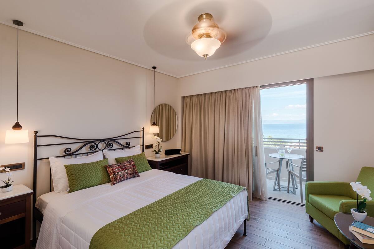 Et hotelværelse med en seng, et natbord, en lampe, en grøn stol og havudsigt fra en balkon.