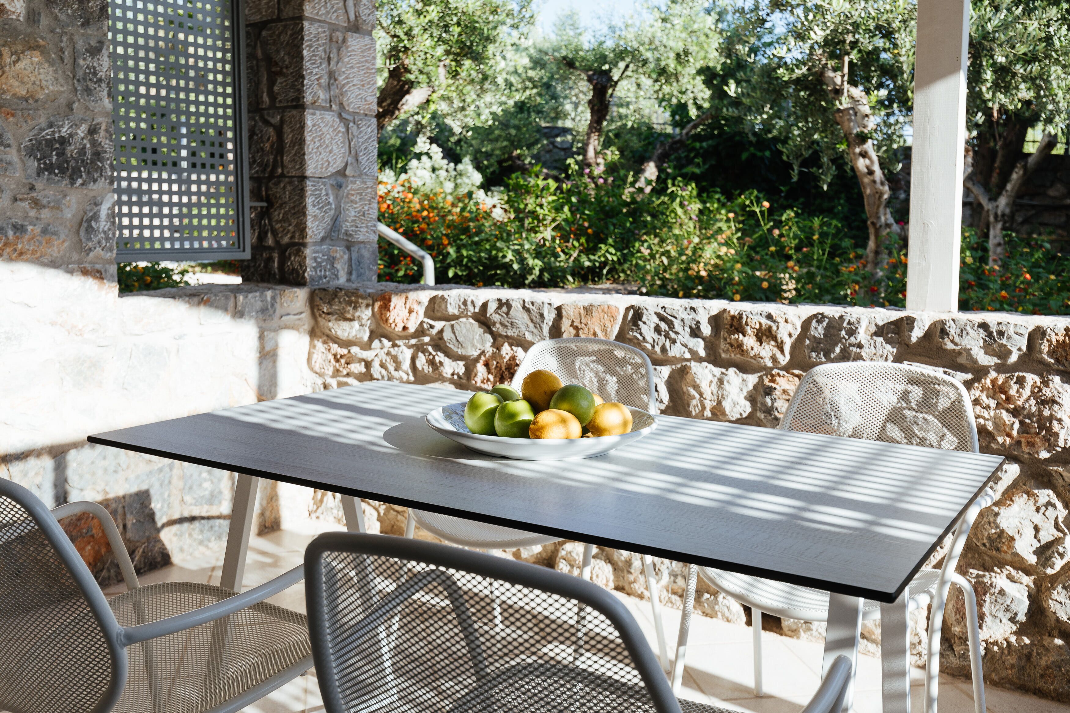 A stone wall with a metal table and chairs, a bowl of fruit, and a view of trees and flowers.