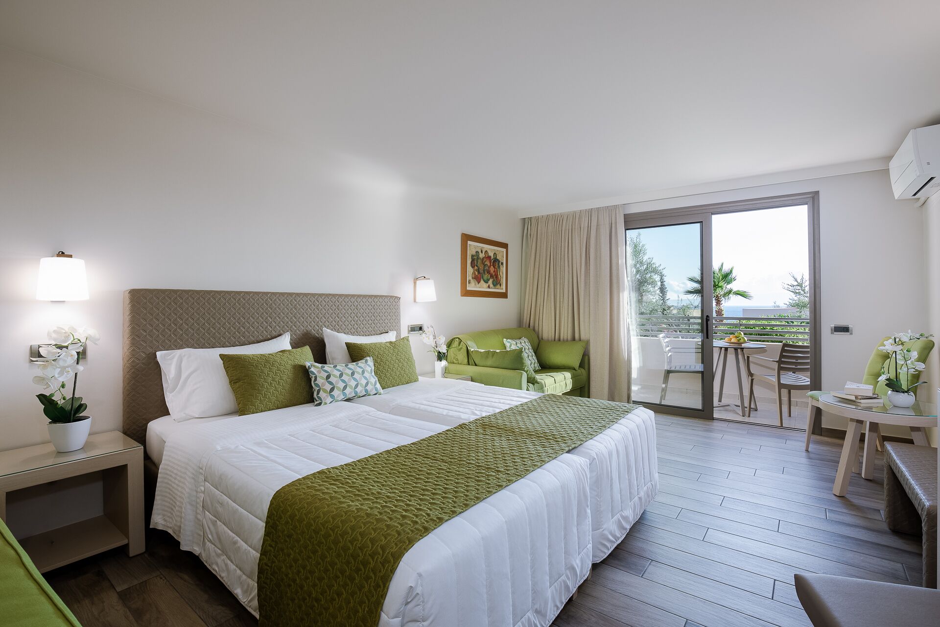 A hotel room with a large bed, a green sofa, a balcony with a table and chairs, and a view of the sea.