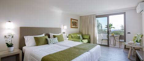 A hotel room with a large bed, a green sofa, a balcony with a table and chairs, and a view of the sea.