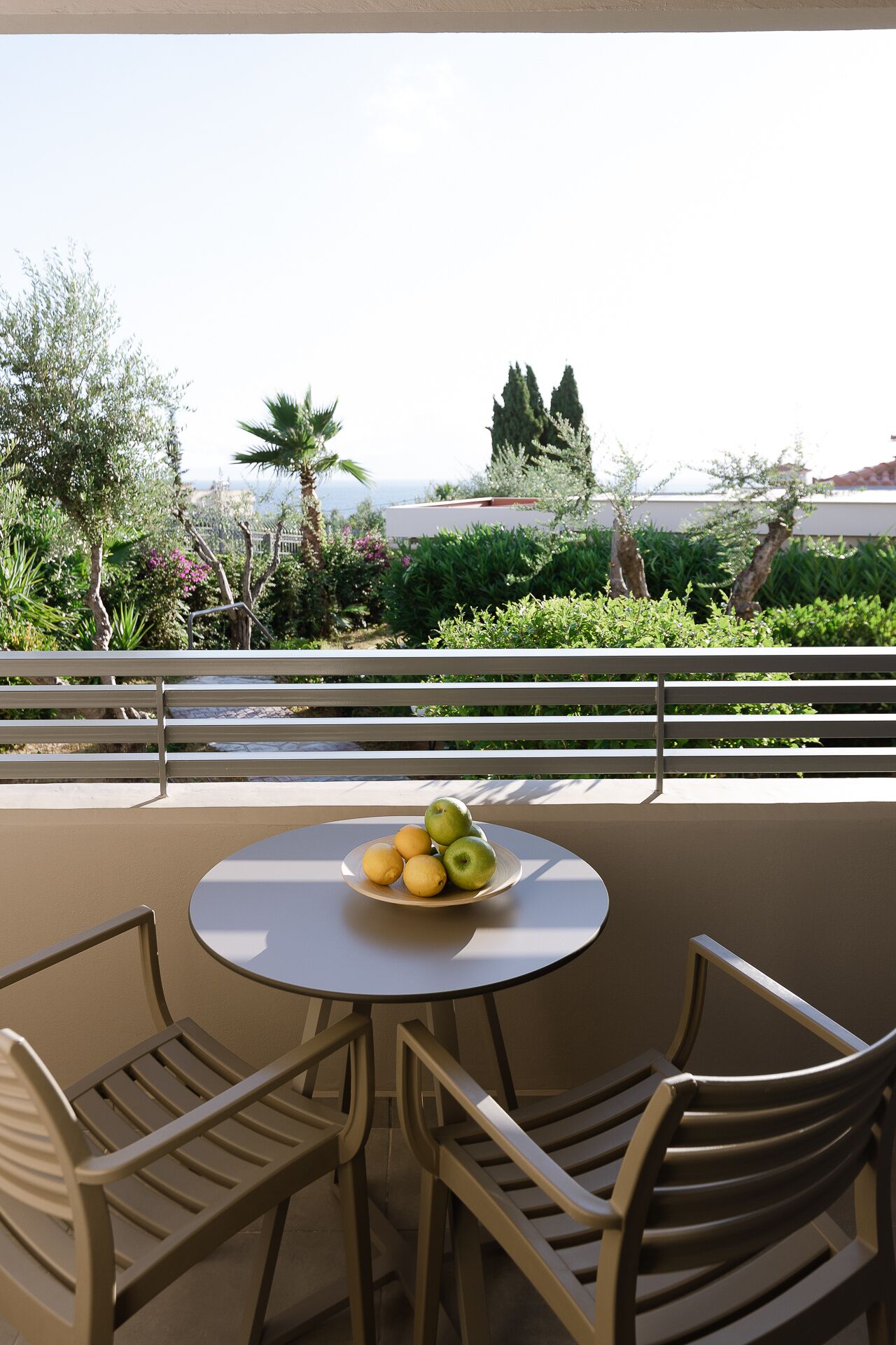 A round table with a fruit bowl on it, surrounded by two chairs, on a balcony with a railing and a view of greenery and buildings.
