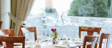 A dining table set with white tableware, a potted plant with pink flowers, and a view of greenery outside.