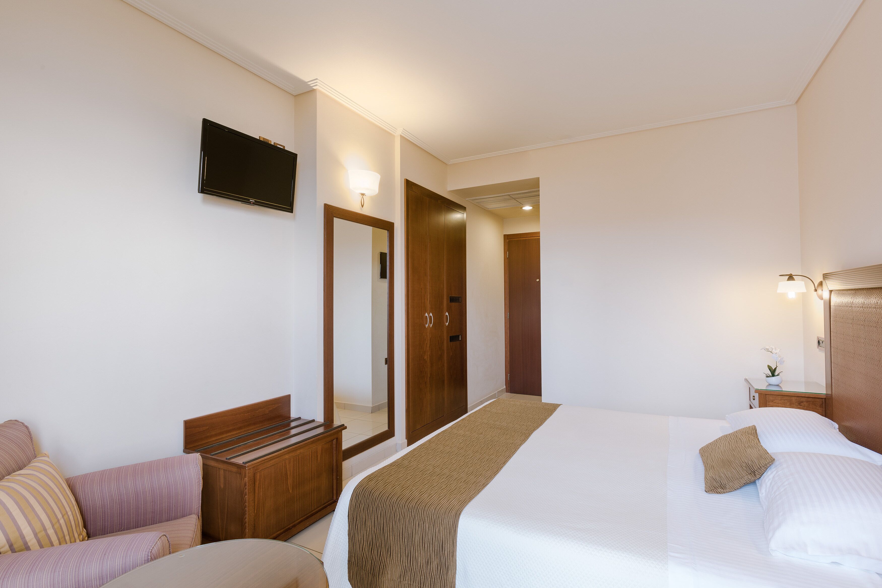 Quarto de hotel com cama, guarda-roupa de madeira, televisão na parede e pequena mesa com abajur.