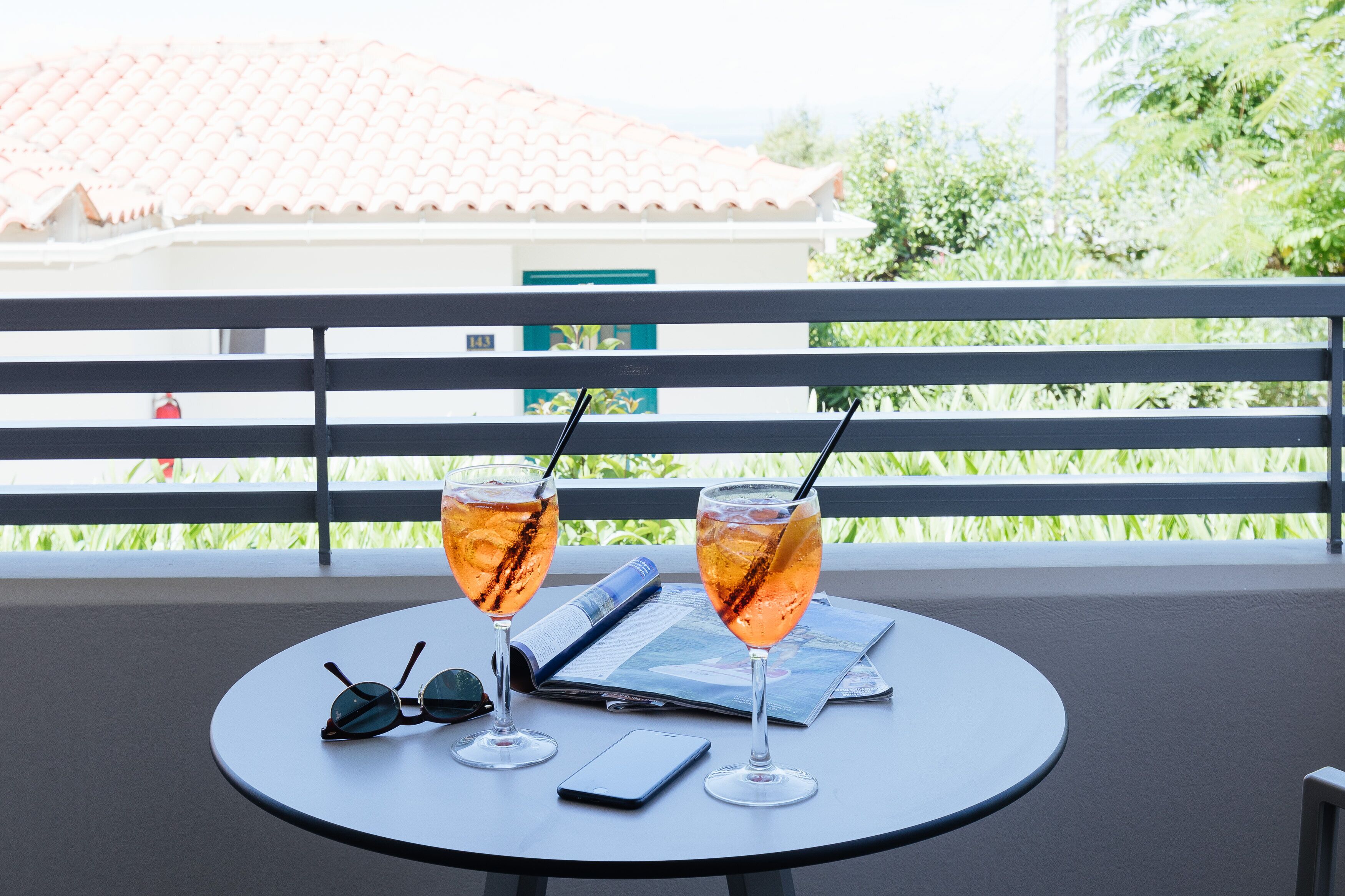 A table with two glasses of a beverage, a pair of sunglasses, and a smartphone on a balcony with a railing and greenery in the background.