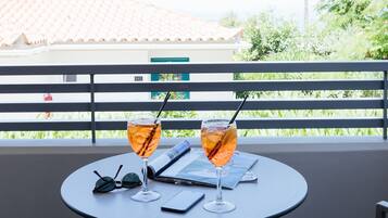 A table with two glasses of a beverage, a pair of sunglasses, and a smartphone on a balcony with a railing and greenery in the background.