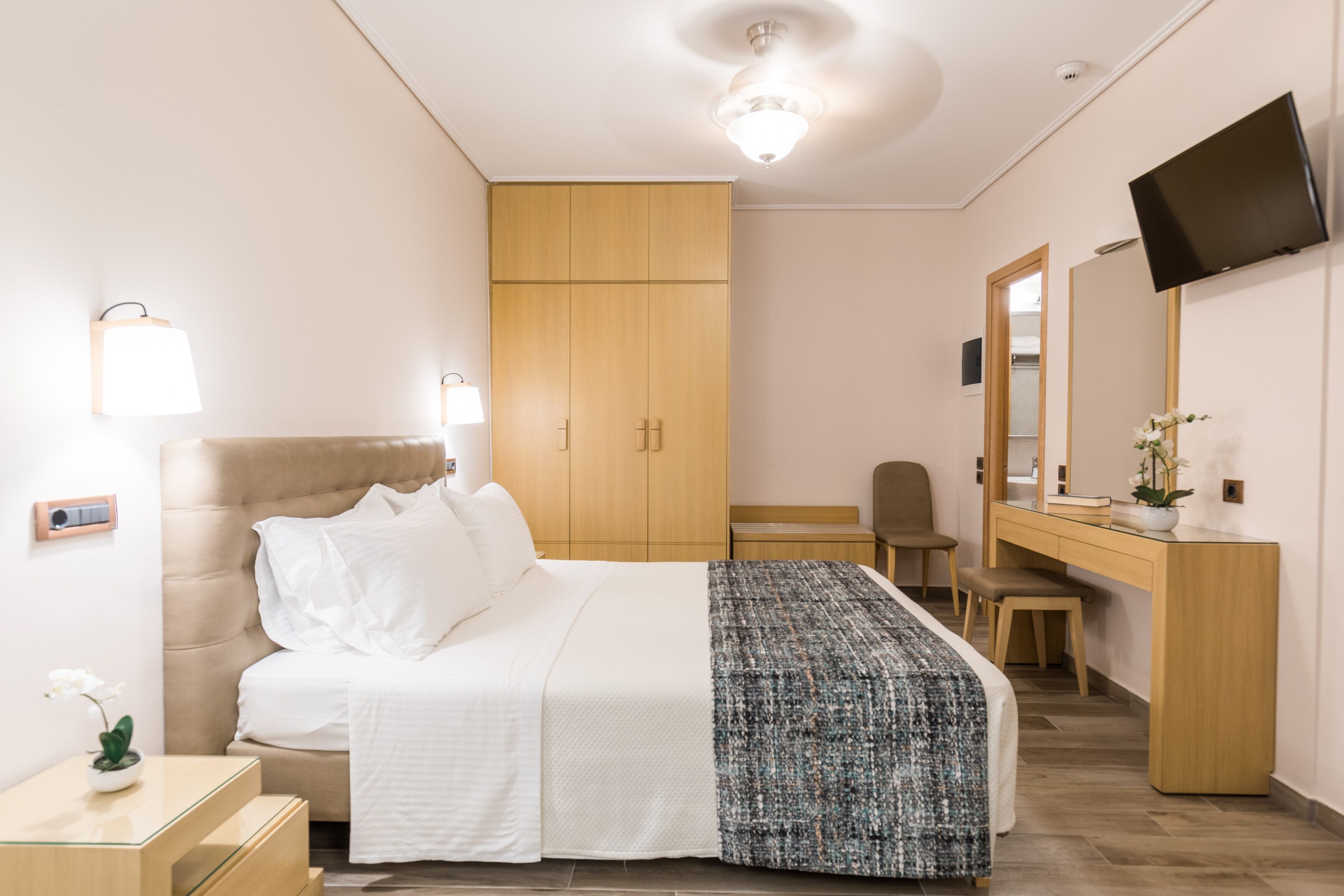 A hotel room with a bed, bedside table, lamp, wardrobe, desk, chair, and a TV mounted on the wall.