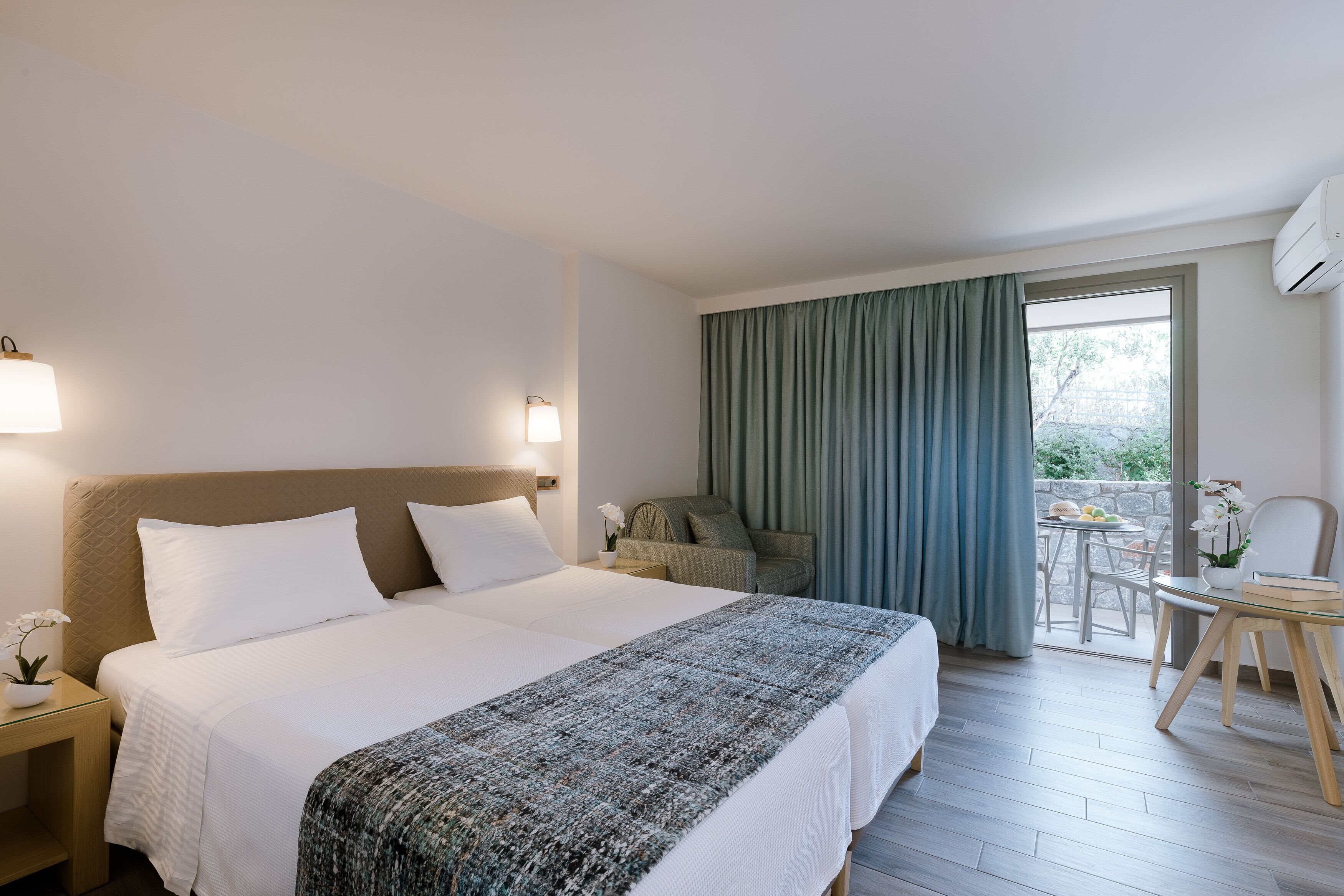 Una habitación de hotel con una cama grande, una mesita con una silla, un balcón con mesa y sillas, y un equipo de aire acondicionado.