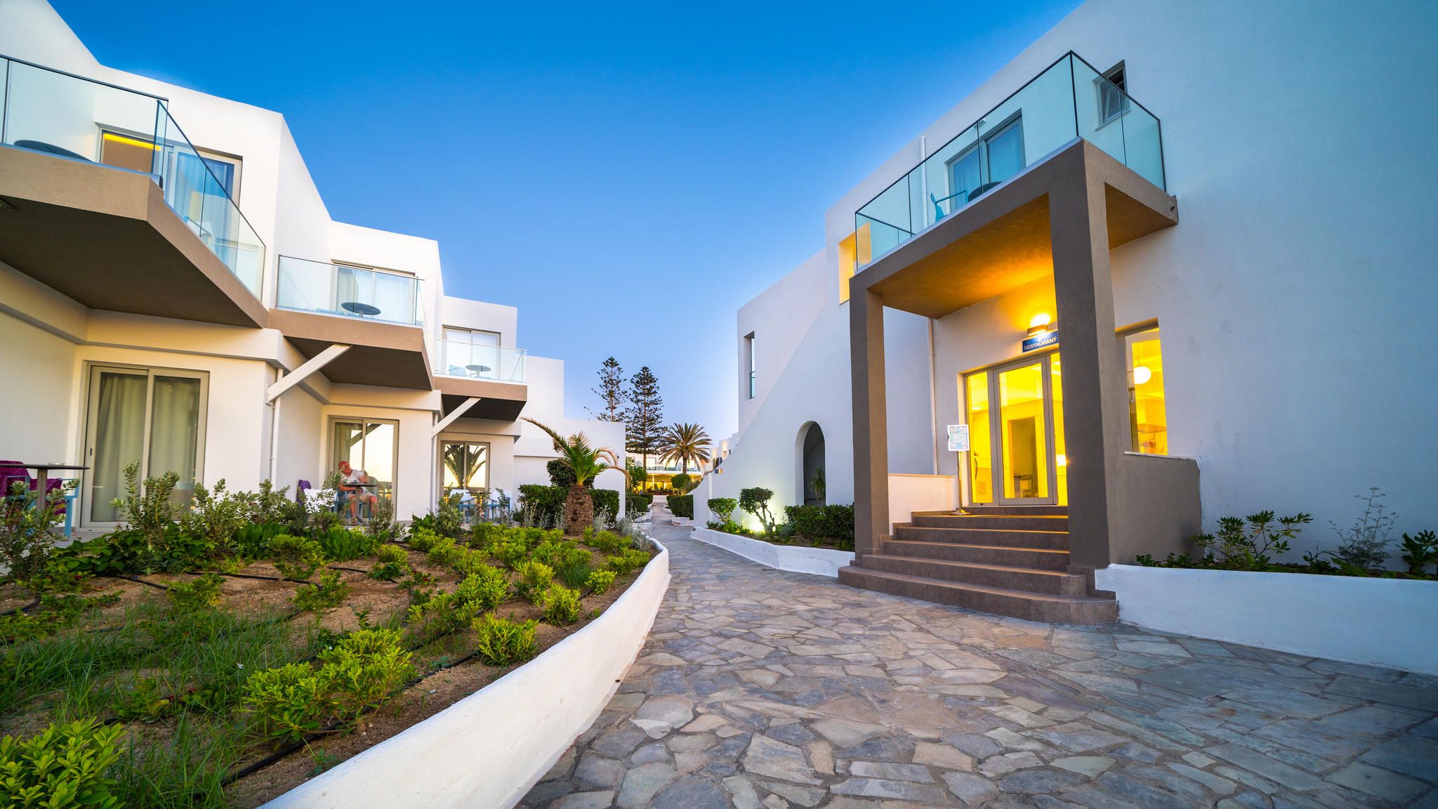 A modern hotel with a stone pathway leading to the entrance, a restaurant sign, and a clear sky.