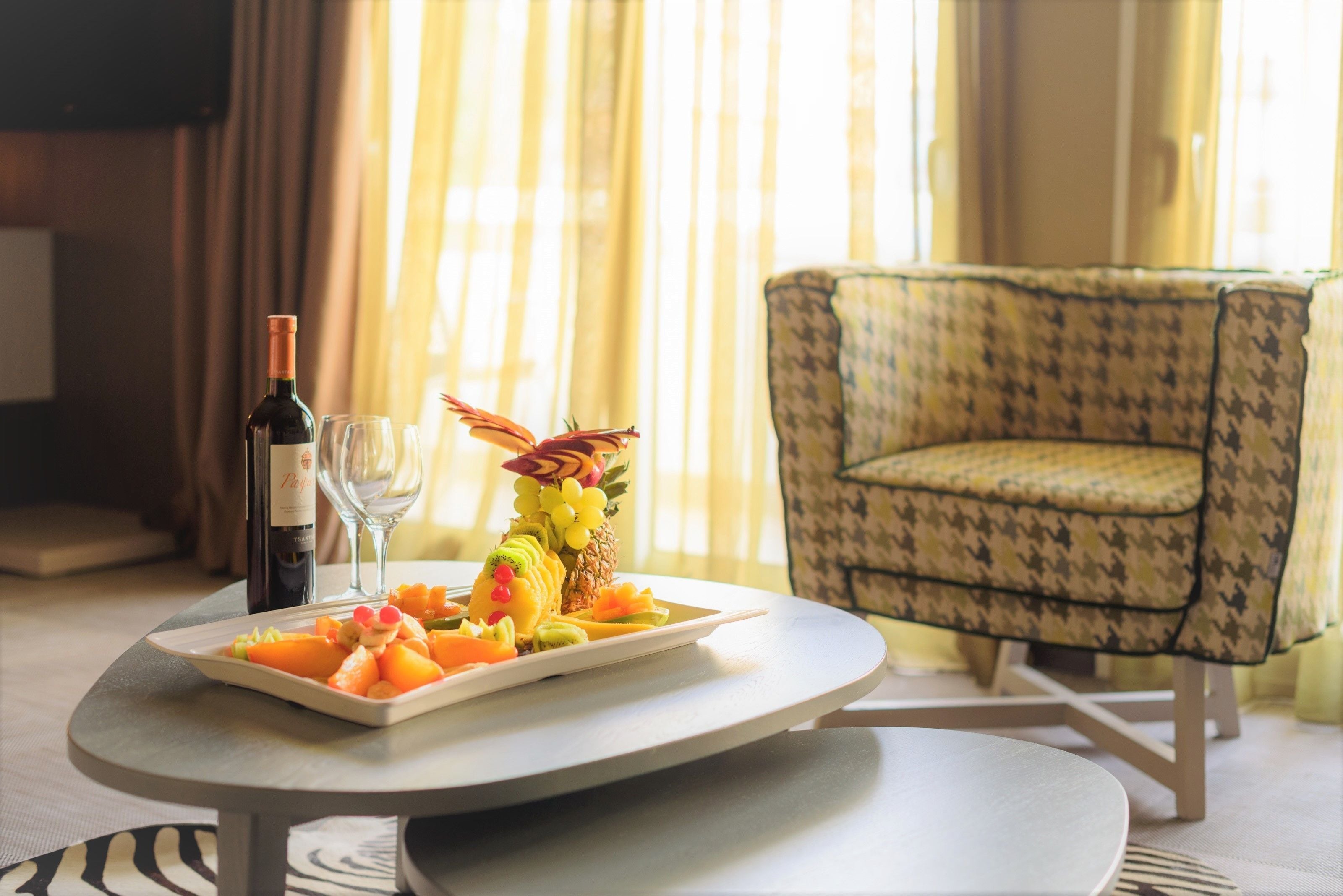 A dining table with a fruit platter, a bottle of wine, and two glasses. There is a patterned armchair in the background.