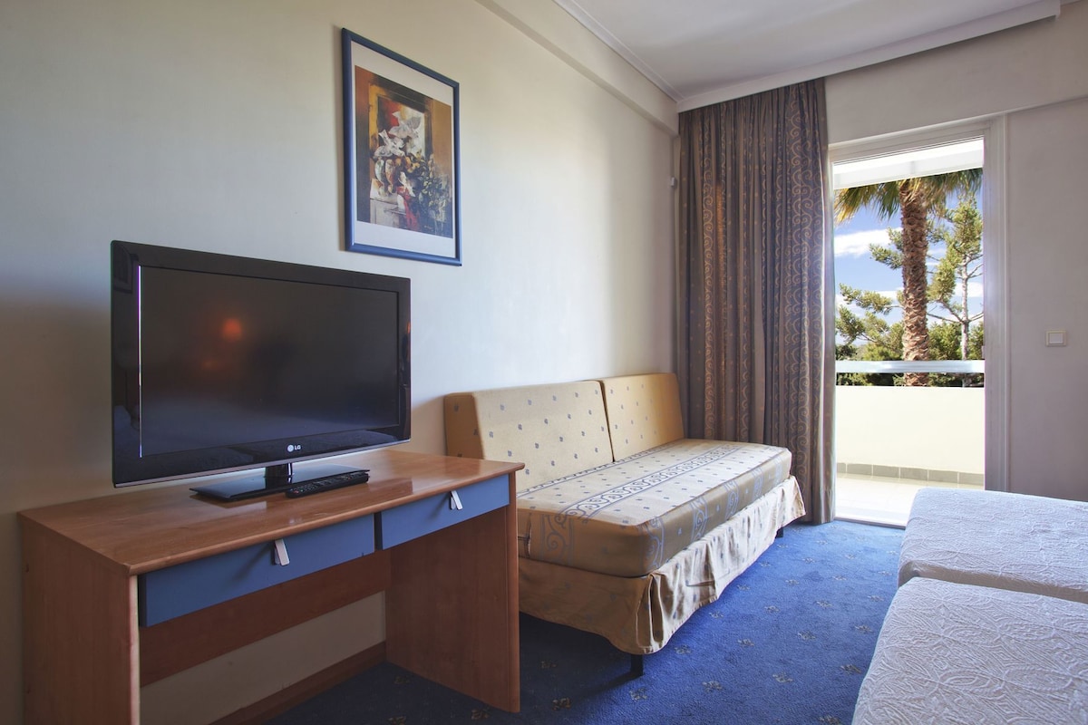 A hotel room with a television on a wooden desk, a sofa, a bed, and a balcony with a view of trees and a building.