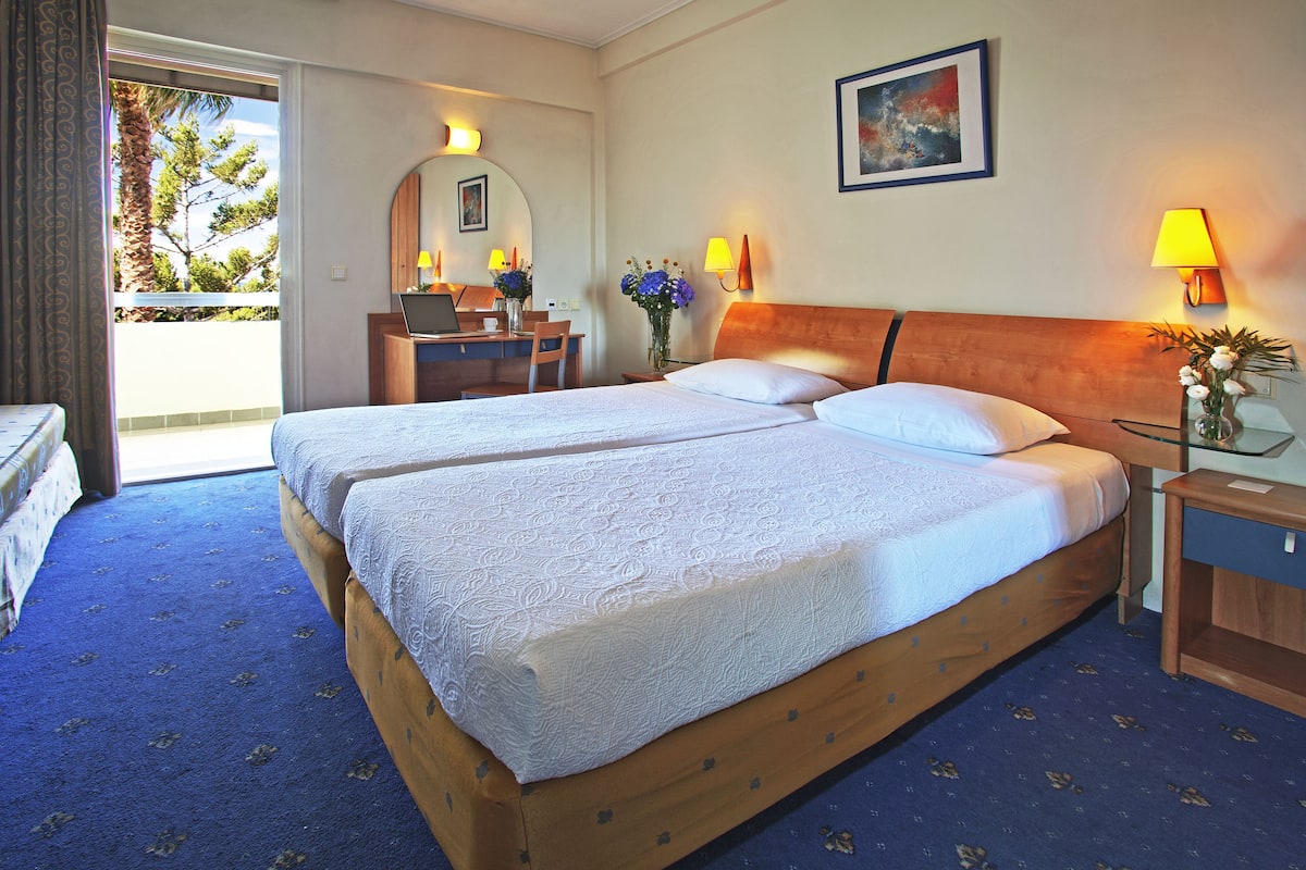 A hotel room with two beds, a desk with a chair, a laptop, a vase with flowers, and a painting on the wall.