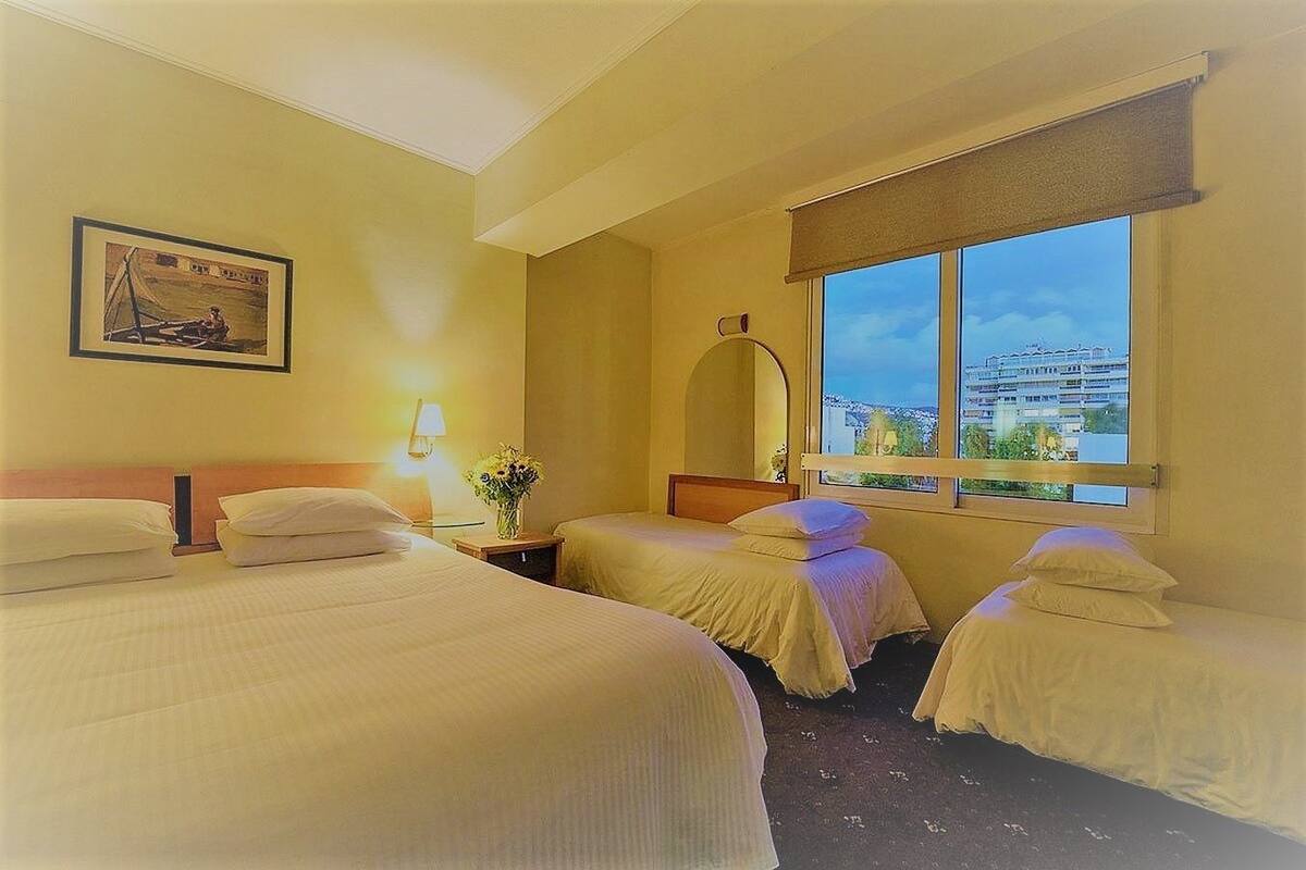 A hotel room with two beds, a nightstand with a lamp, a window with a view of buildings, and a framed picture on the wall.