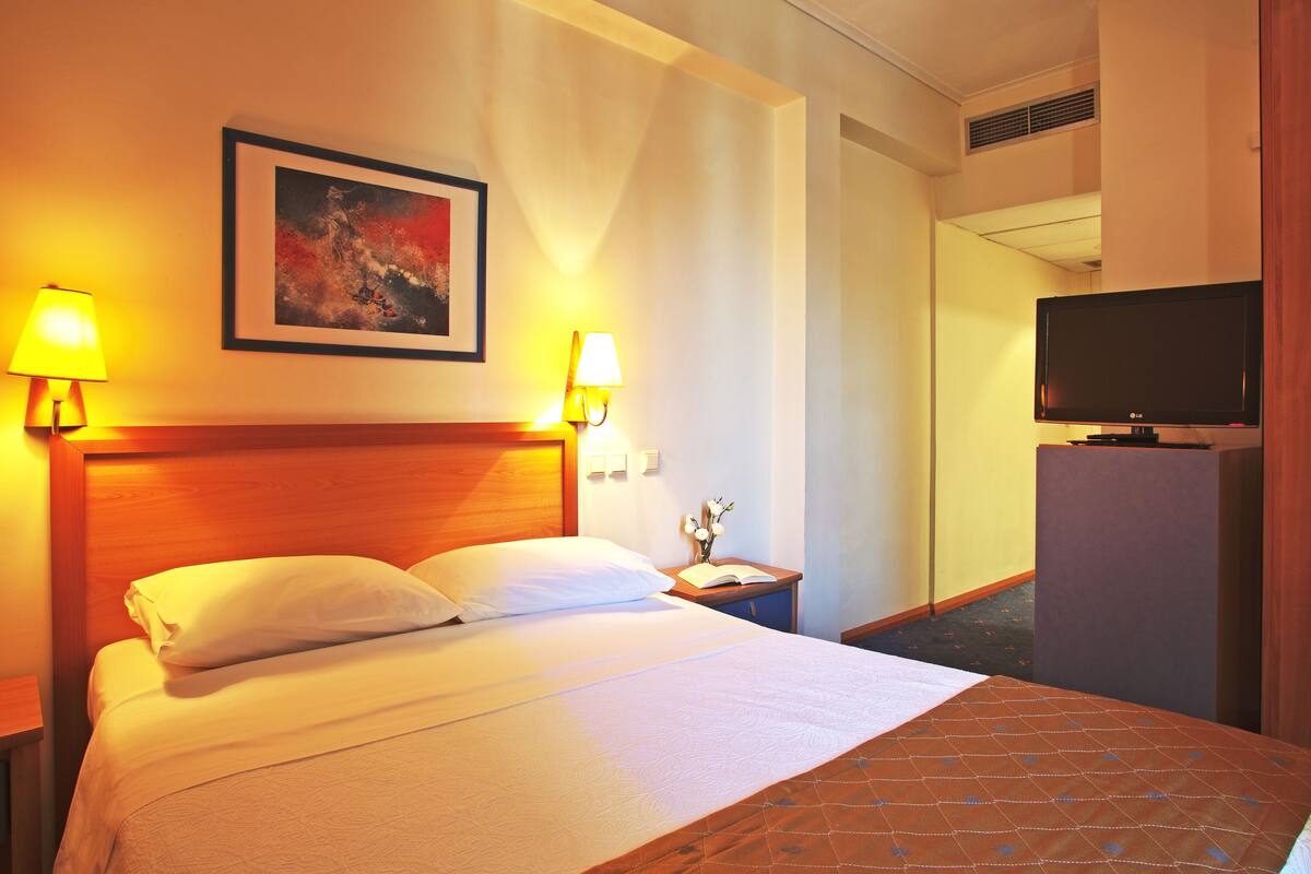 A hotel room with a bed, two bedside lamps, a painting, a television, and a desk with a lamp.