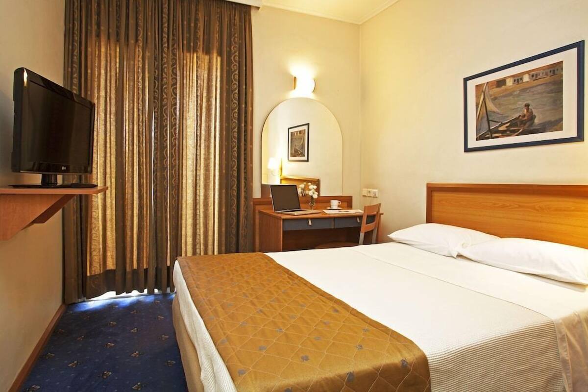 A hotel room with a bed, a desk with a laptop, a television, and a painting on the wall.