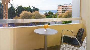 Un balcón con mesa y sillas, un vaso de zumo de naranja y vista al mar y edificios.